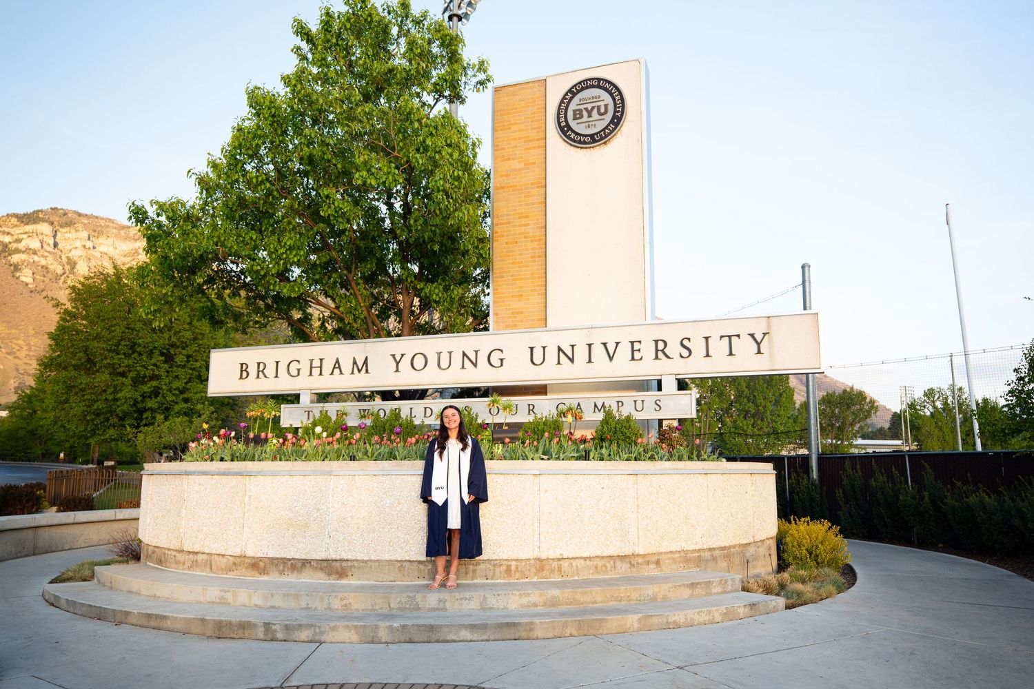 Woman posing in front of Brigham Young University campus sign surrounded by trees and mountains at golden hour.