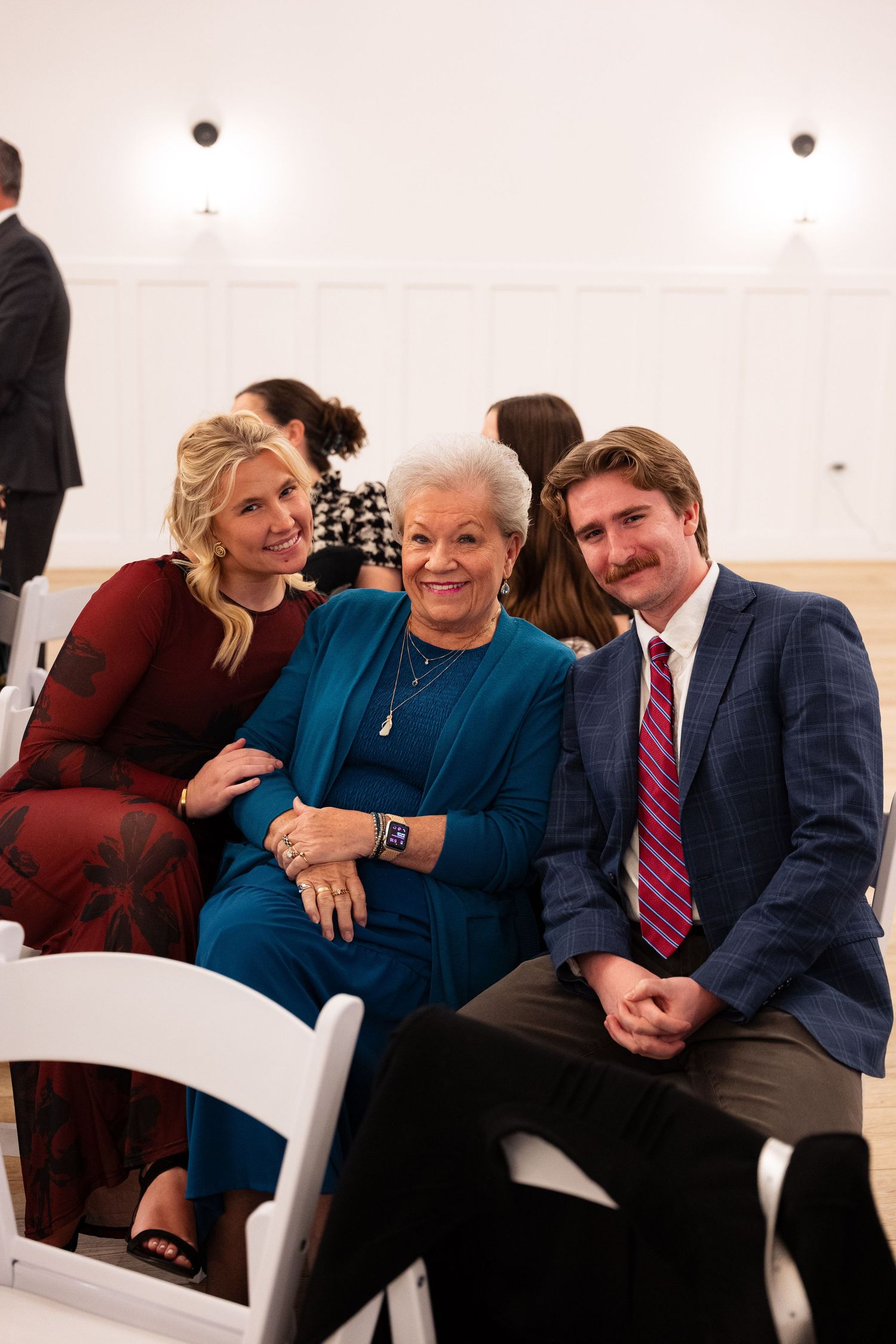 Three wedding guests smile for a photo, two women and a man in formal attire seated in white chairs.