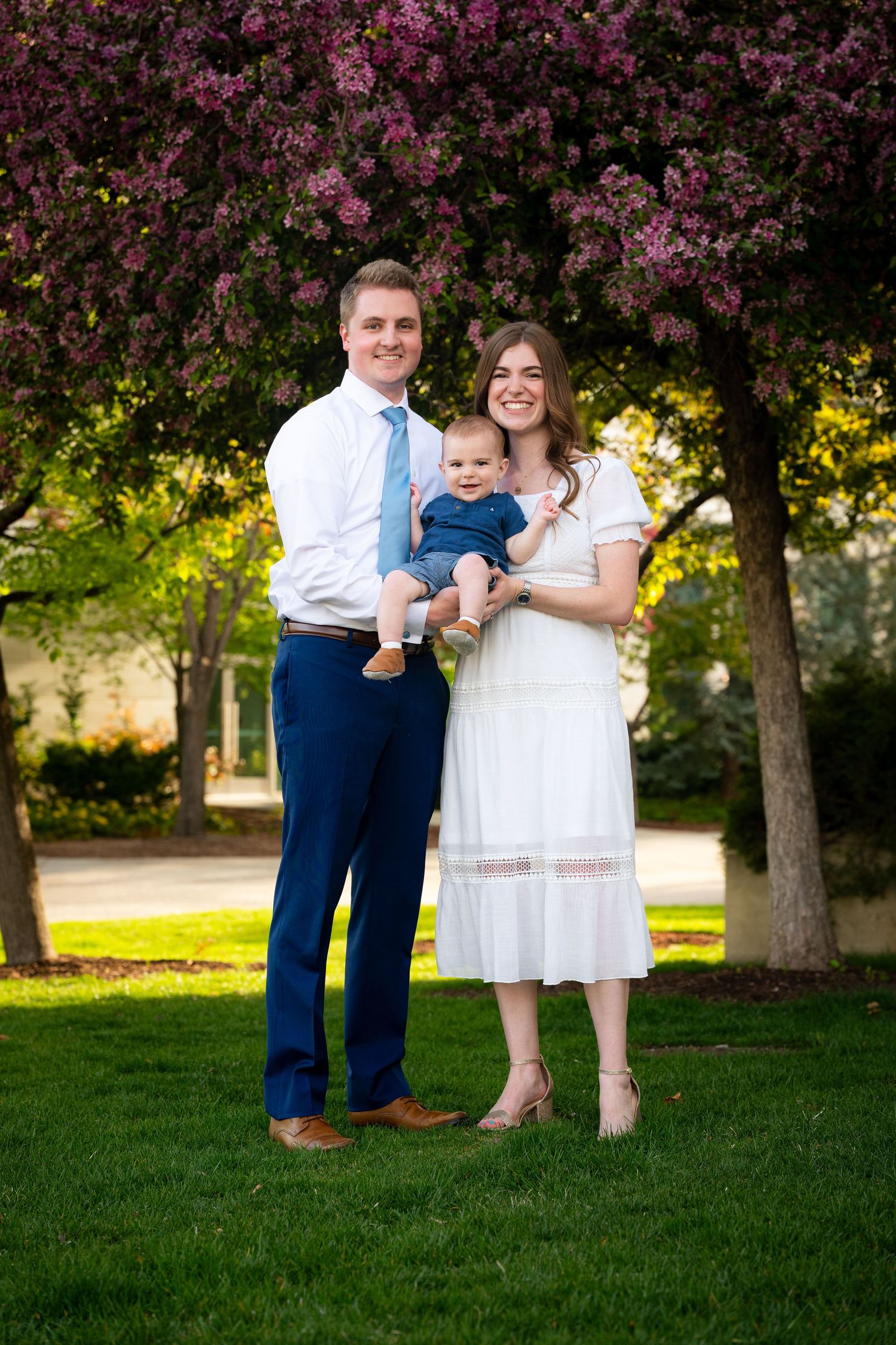 A family of three, including a young child, stands in a park with blooming trees in the background.