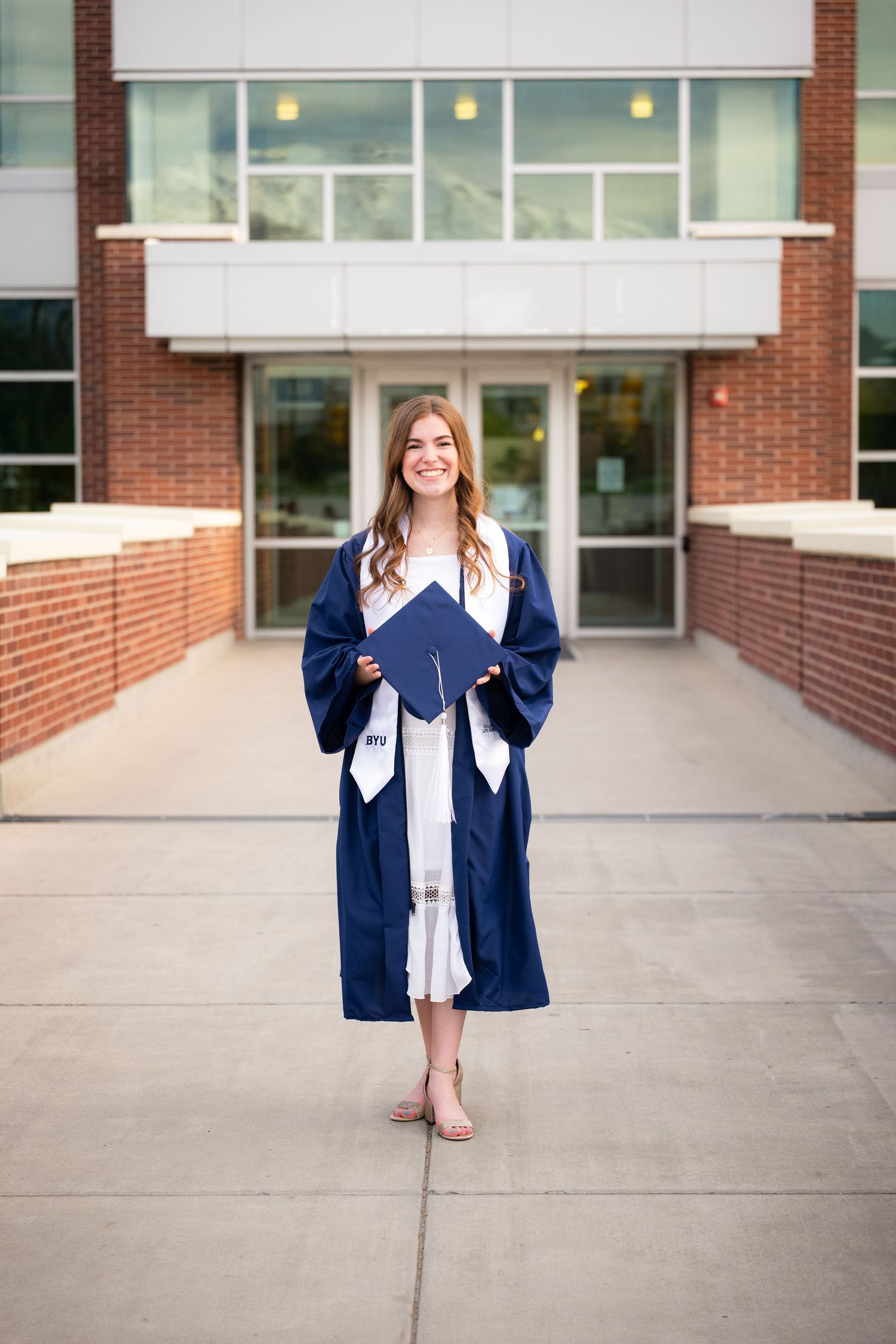 A smiling woman in a graduation gown and cap stands in front of a university building.