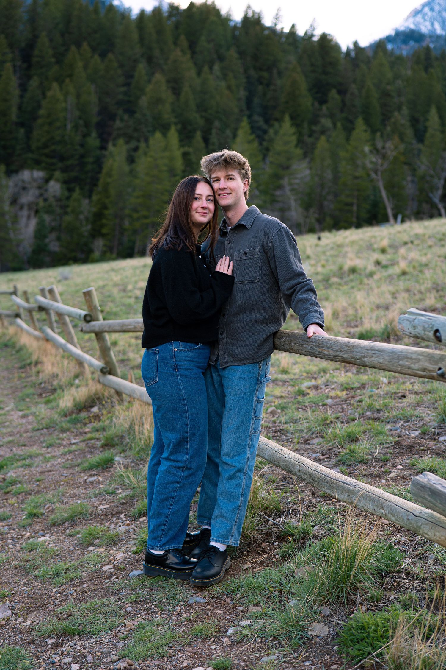 Young couple smiling and embracing by a rustic wooden fence in a scenic mountain meadow with evergreen trees.