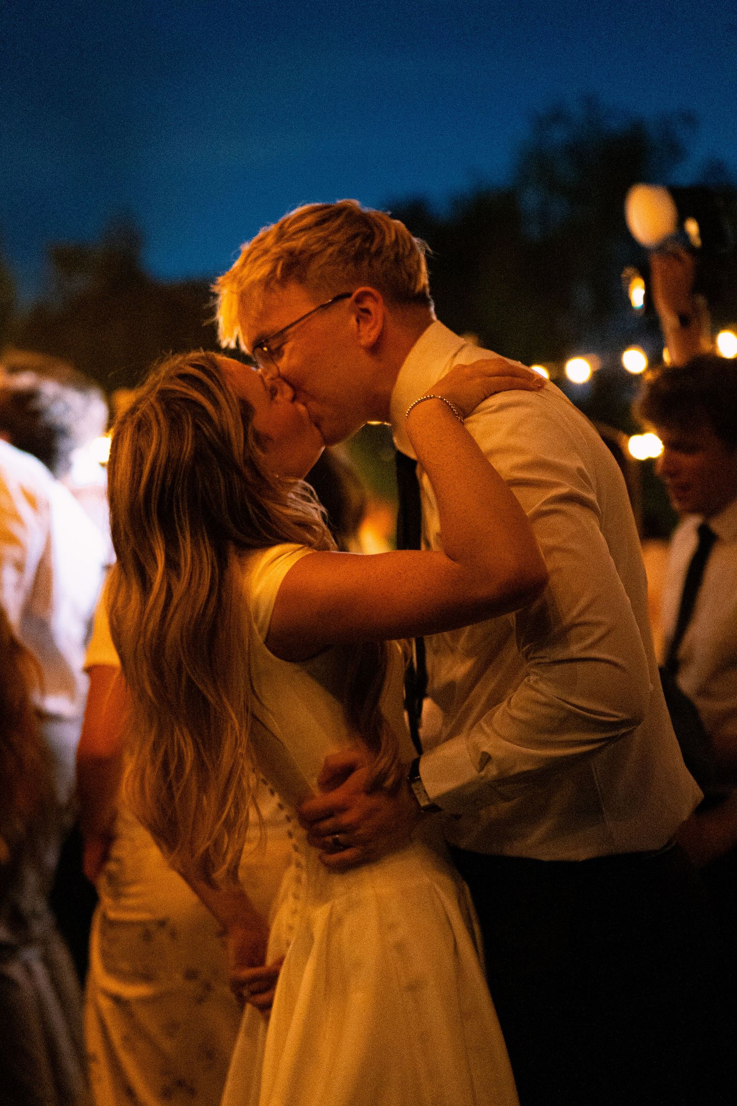 Couple sharing a romantic kiss at an outdoor evening wedding reception under warm string lights.