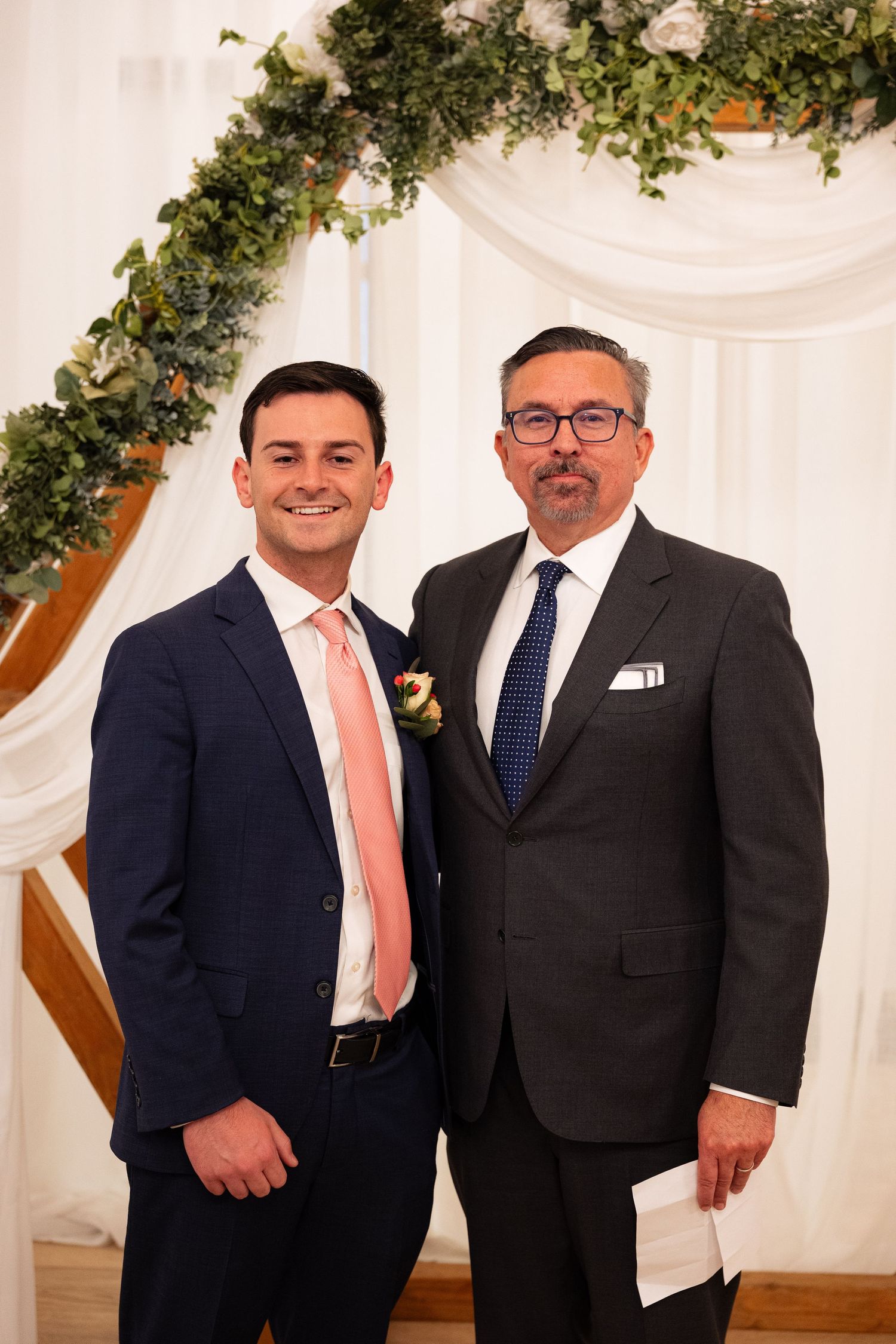 Two men in suits pose together at a wedding ceremony beneath a floral arch with greenery.