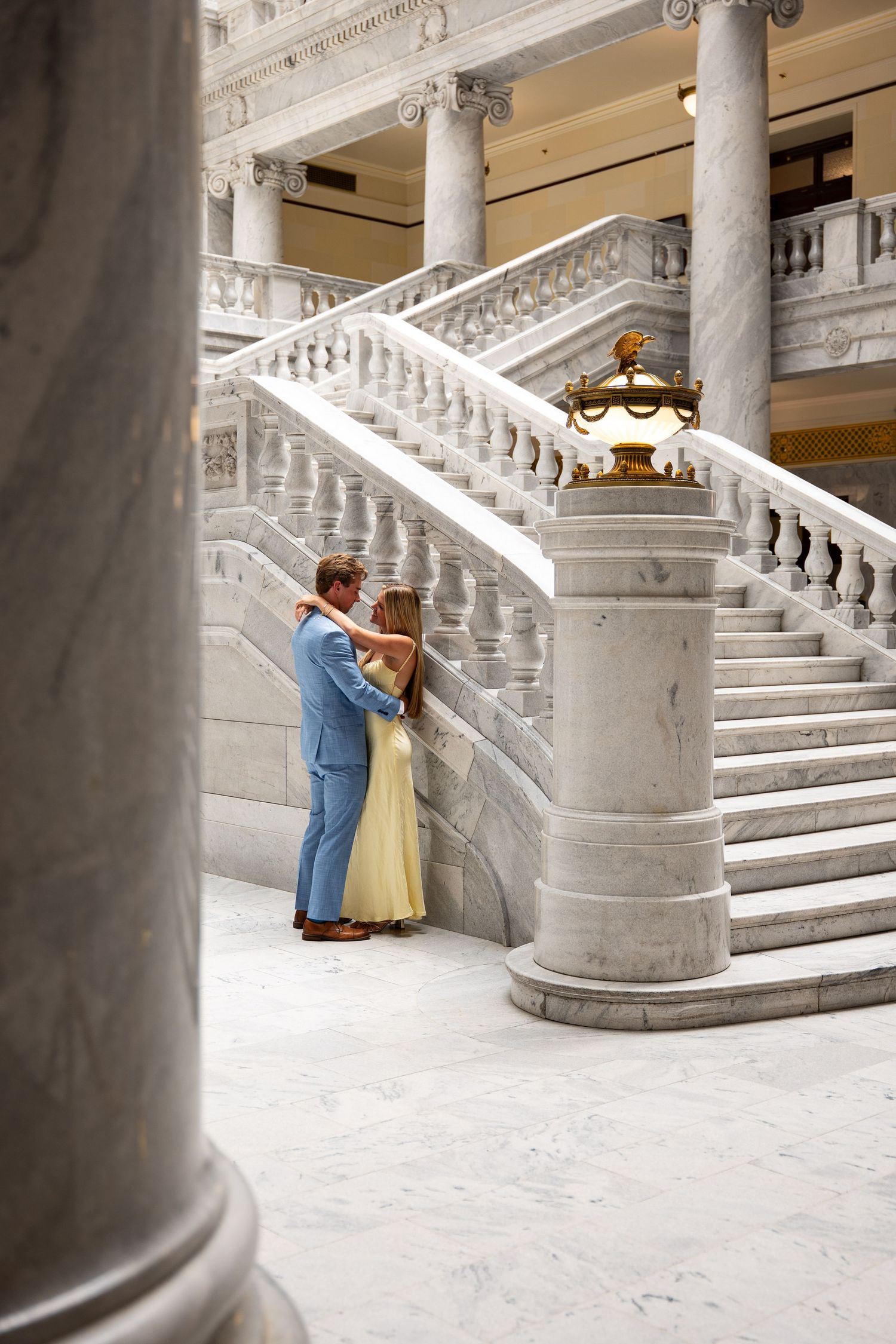 Couple embracing beside marble staircase with ornate balustrade in elegant neoclassical building interior.