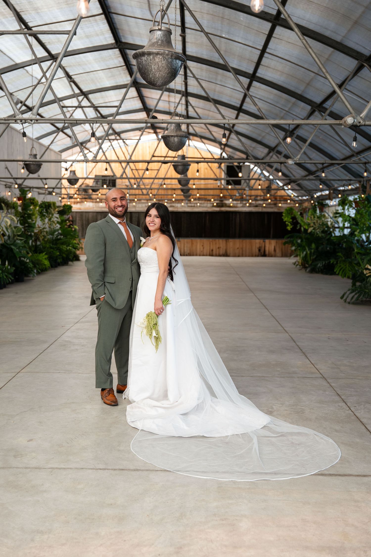Bride and groom pose elegantly in a greenhouse wedding venue with string lights and lush greenery.