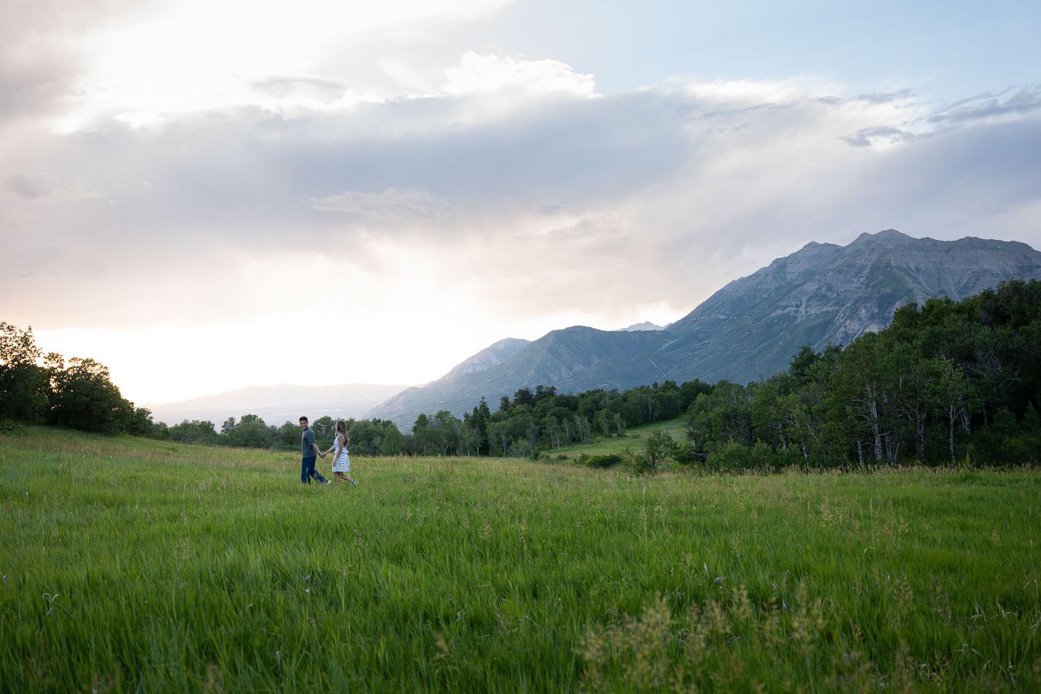 Couple walking through lush green meadow with mountain backdrop at sunset.