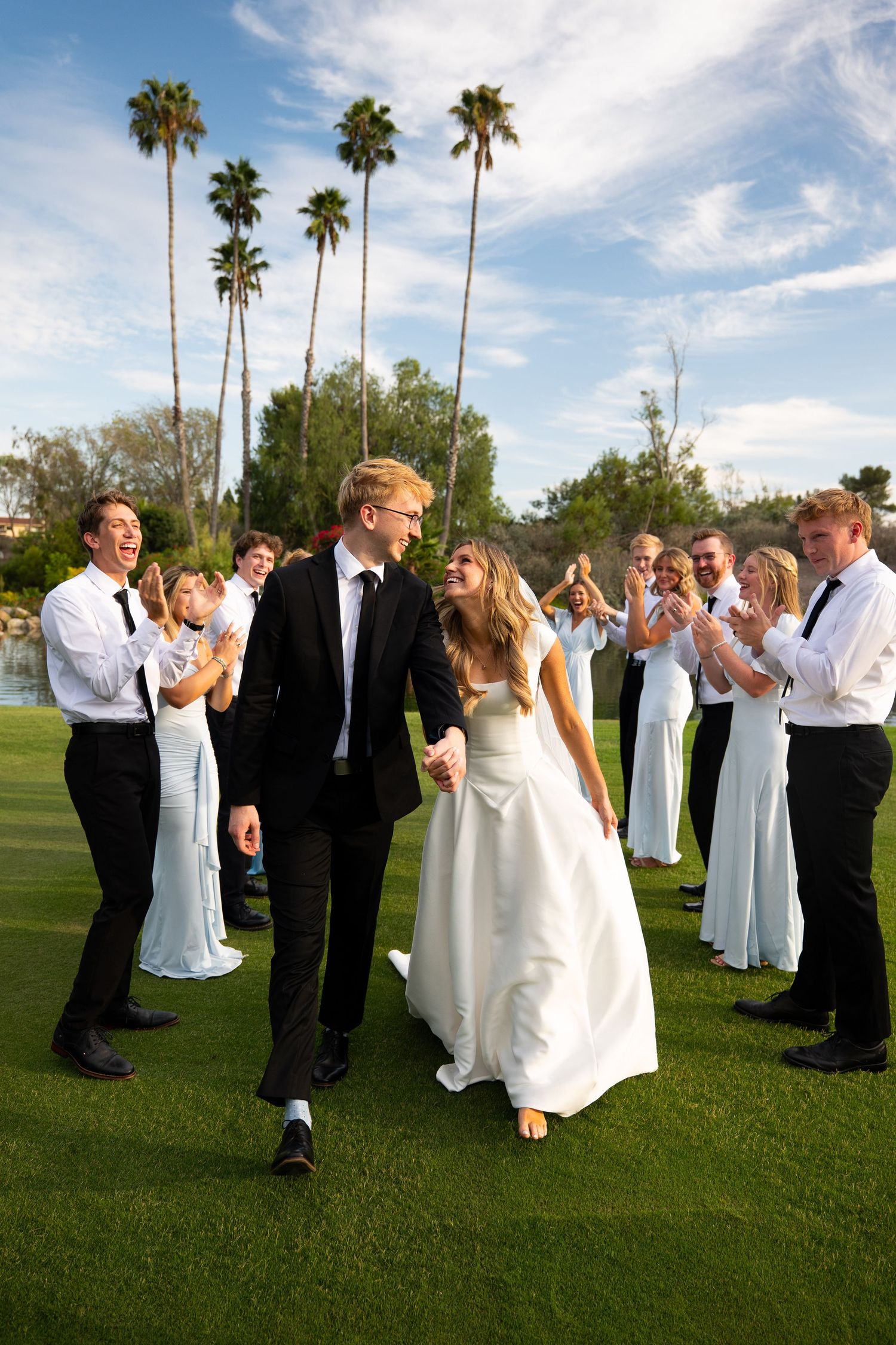 Bride and groom walk hand-in-hand through cheering guests on green lawn with palm trees at outdoor wedding.