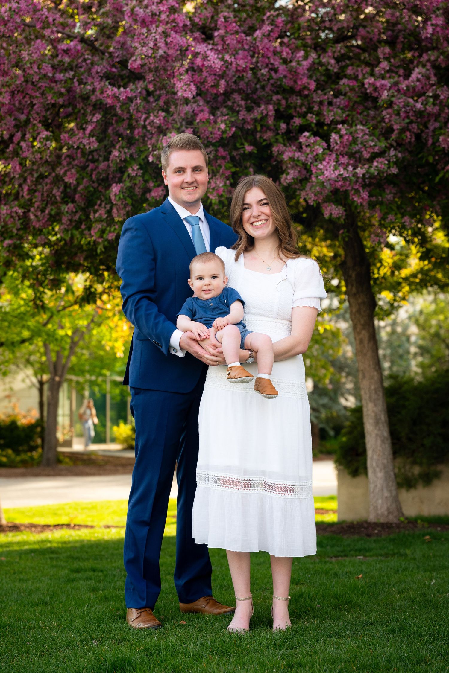 A family of three, including a young child, stands in a park with blooming trees in the background.