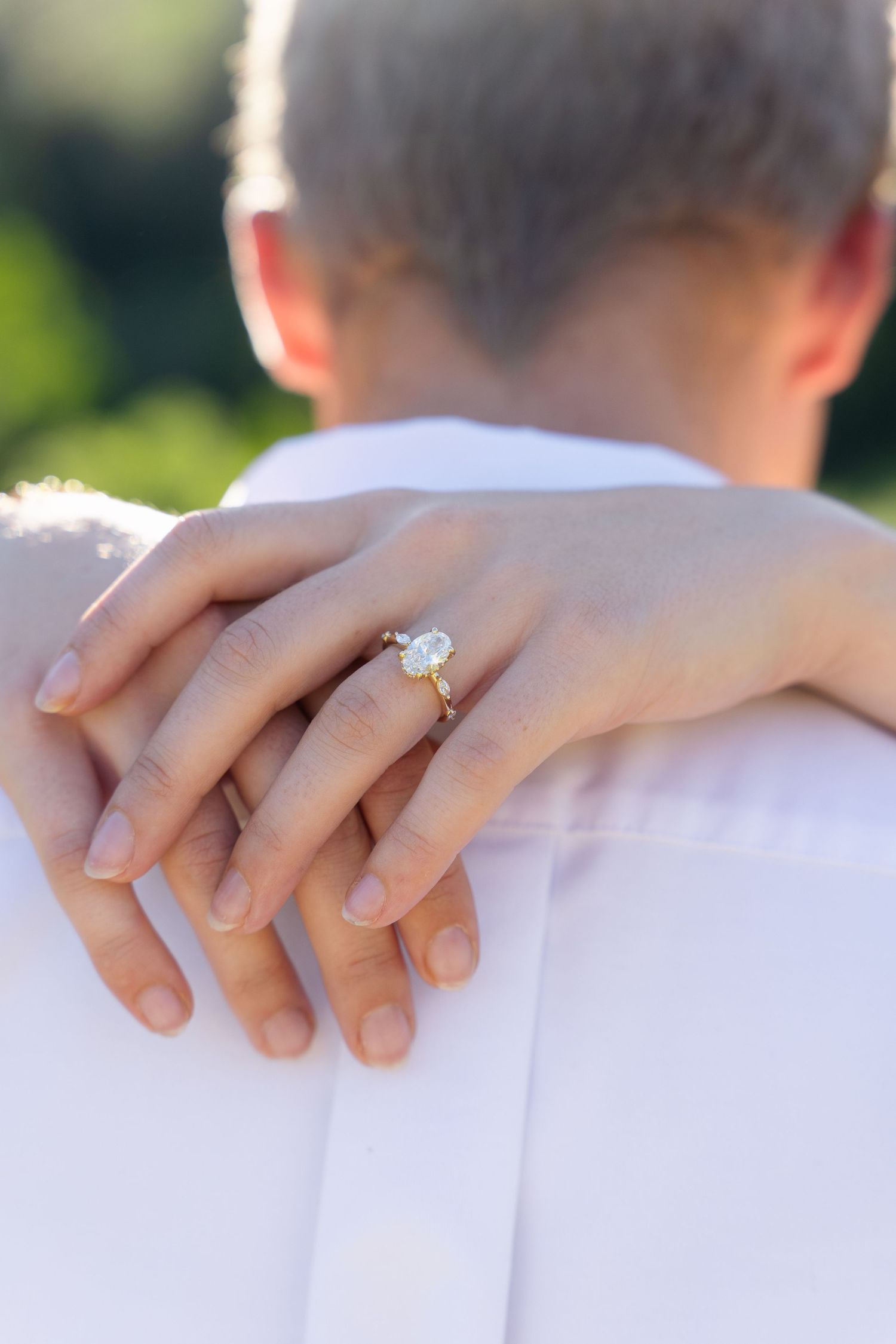Close-up of a woman's hand with a diamond engagement ring resting on a man's shoulder in an outdoor setting.