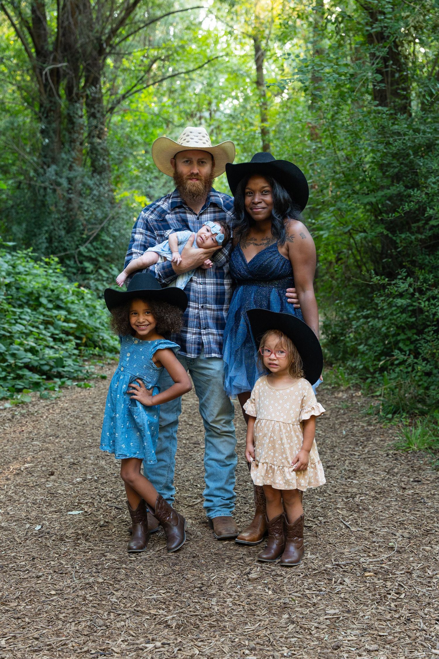 Family of five in cowboy hats posing on a forest path, parents holding newborn with two young girls in boots.