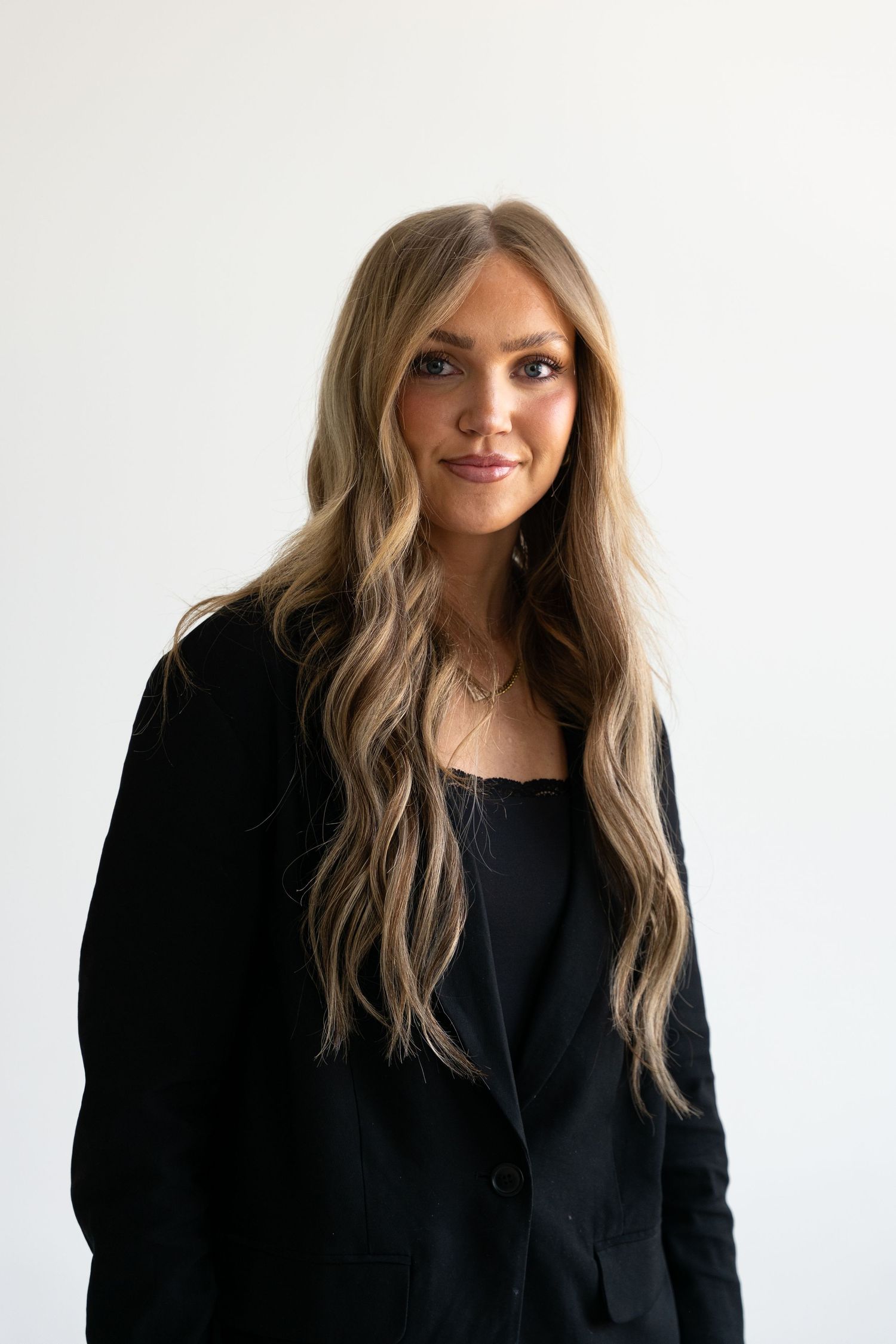Woman with long wavy blonde hair smiling softly in a black blazer and cami against a white background.