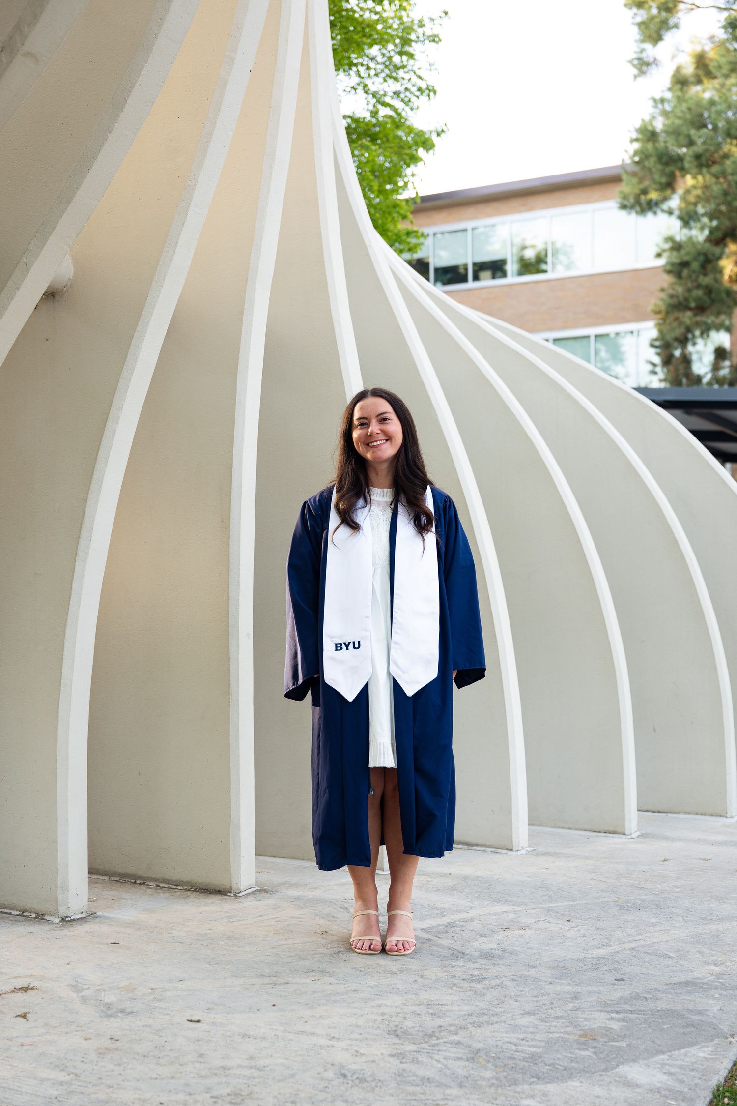 Young woman in navy blue BYU graduation gown and white stole smiling outdoors near architectural arches.