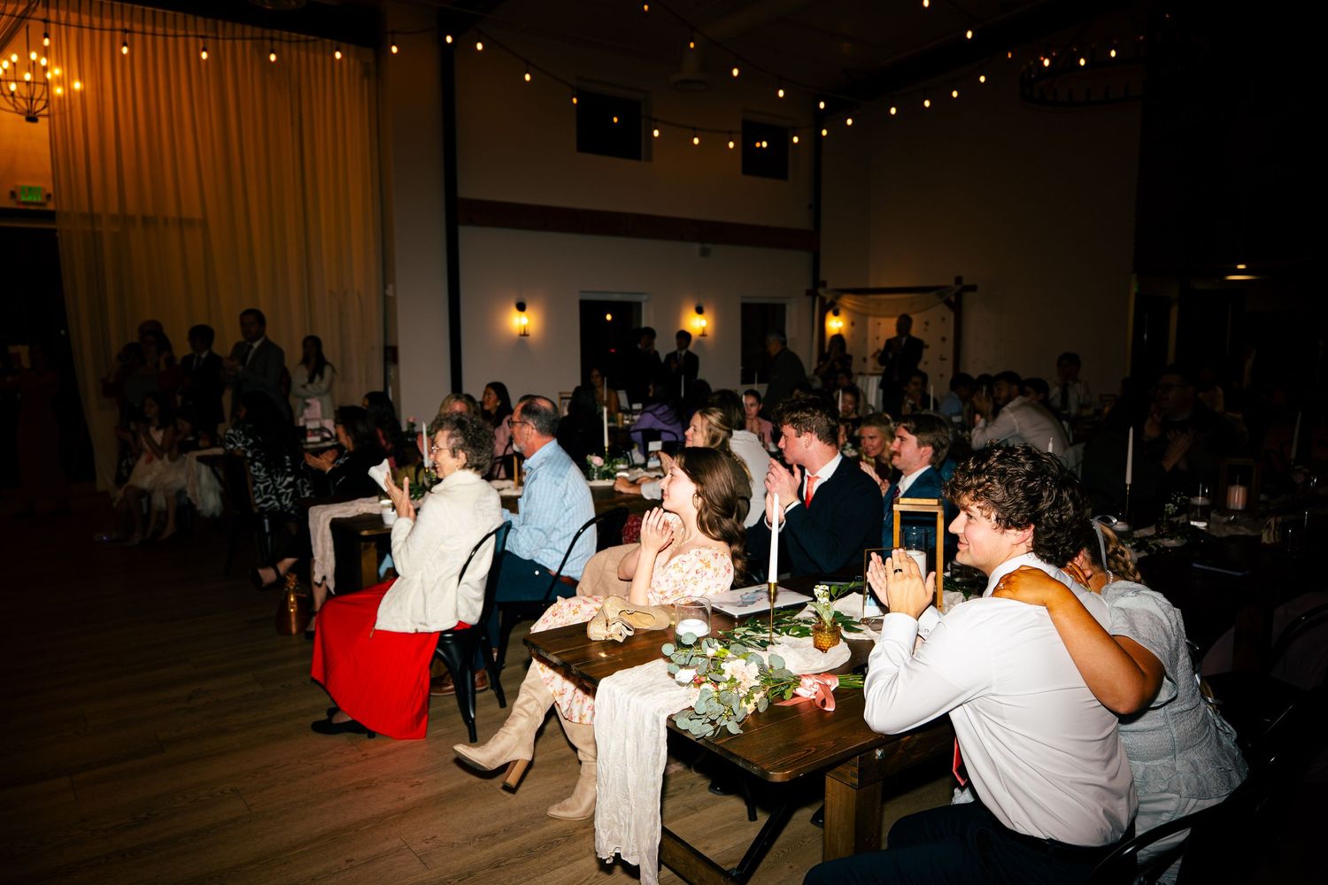 Wedding reception guests seated at long wooden tables under warm string lights.