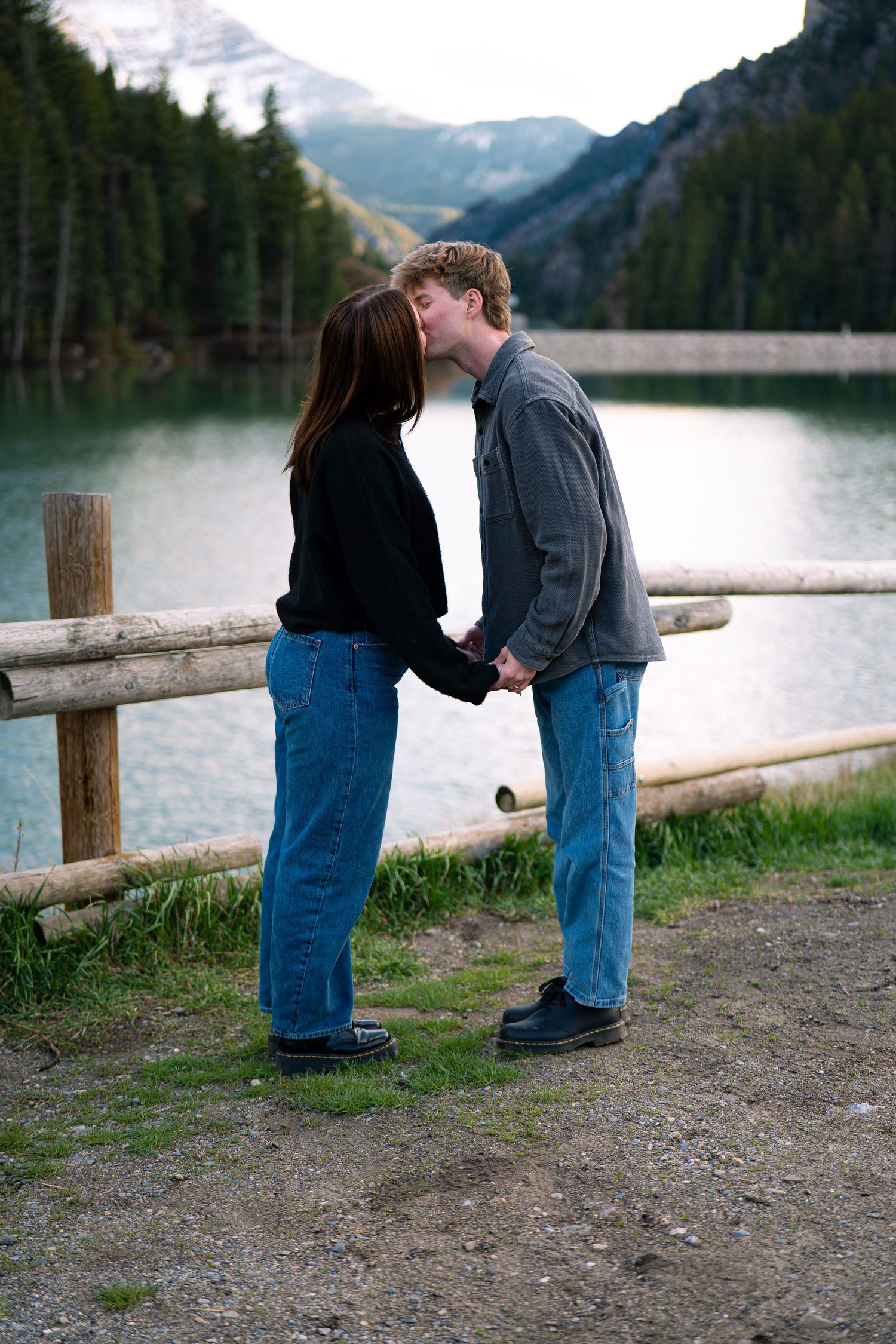 Couple kissing and holding hands by a wooden fence with a mountain lake and forest backdrop.