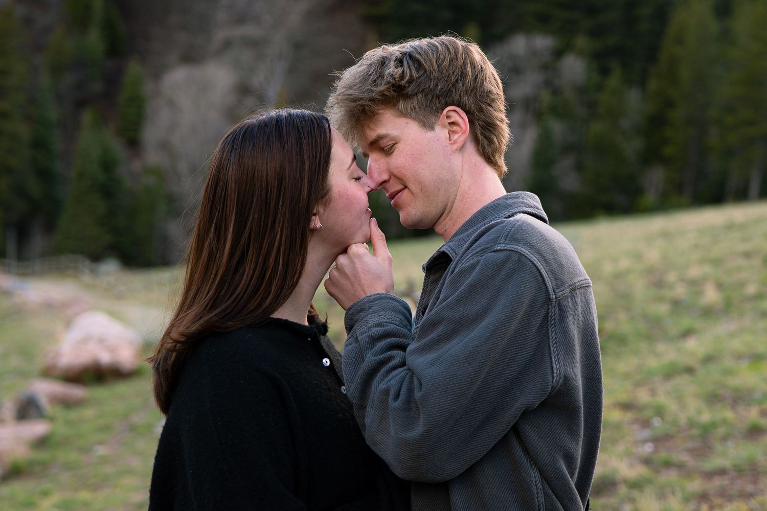 A young couple shares a romantic moment outdoors, with the man gently touching the woman's chin near a rocky landscape.