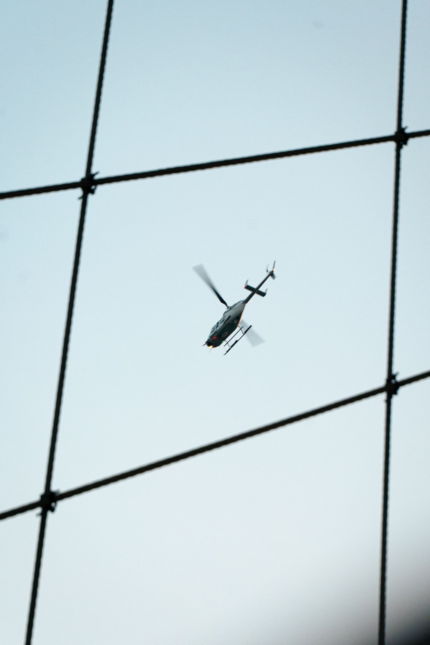 Close-up view of a helicopter flying over the city, framed by the Brooklyn Bridge's cables.