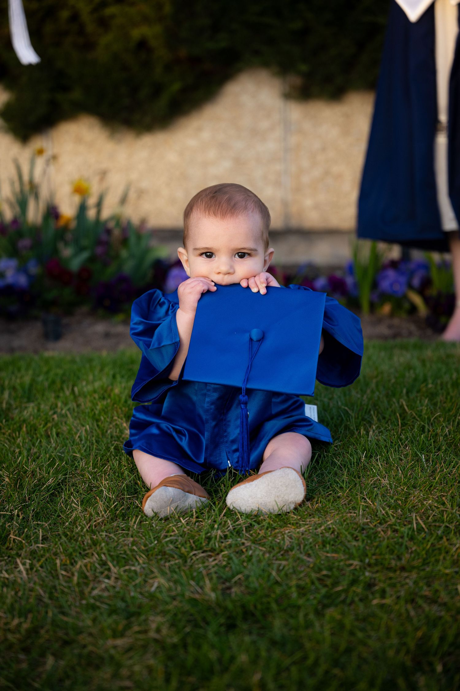 A baby boy sits on grass wearing a blue outfit and looking at the camera.