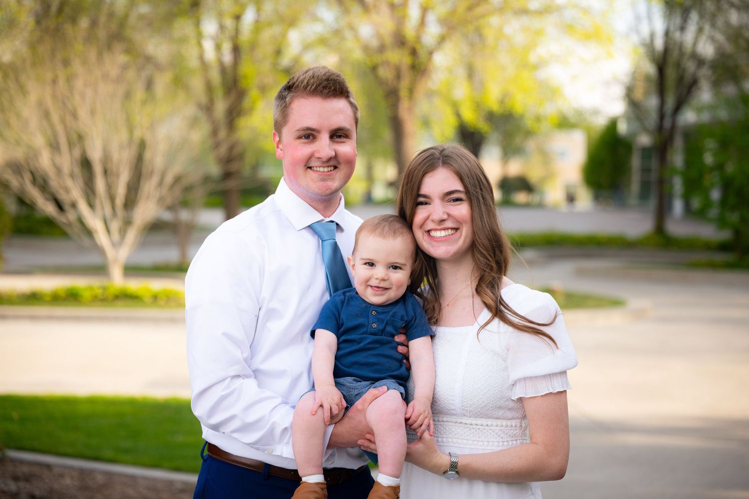 A family of three, including a baby, pose for a portrait outdoors.