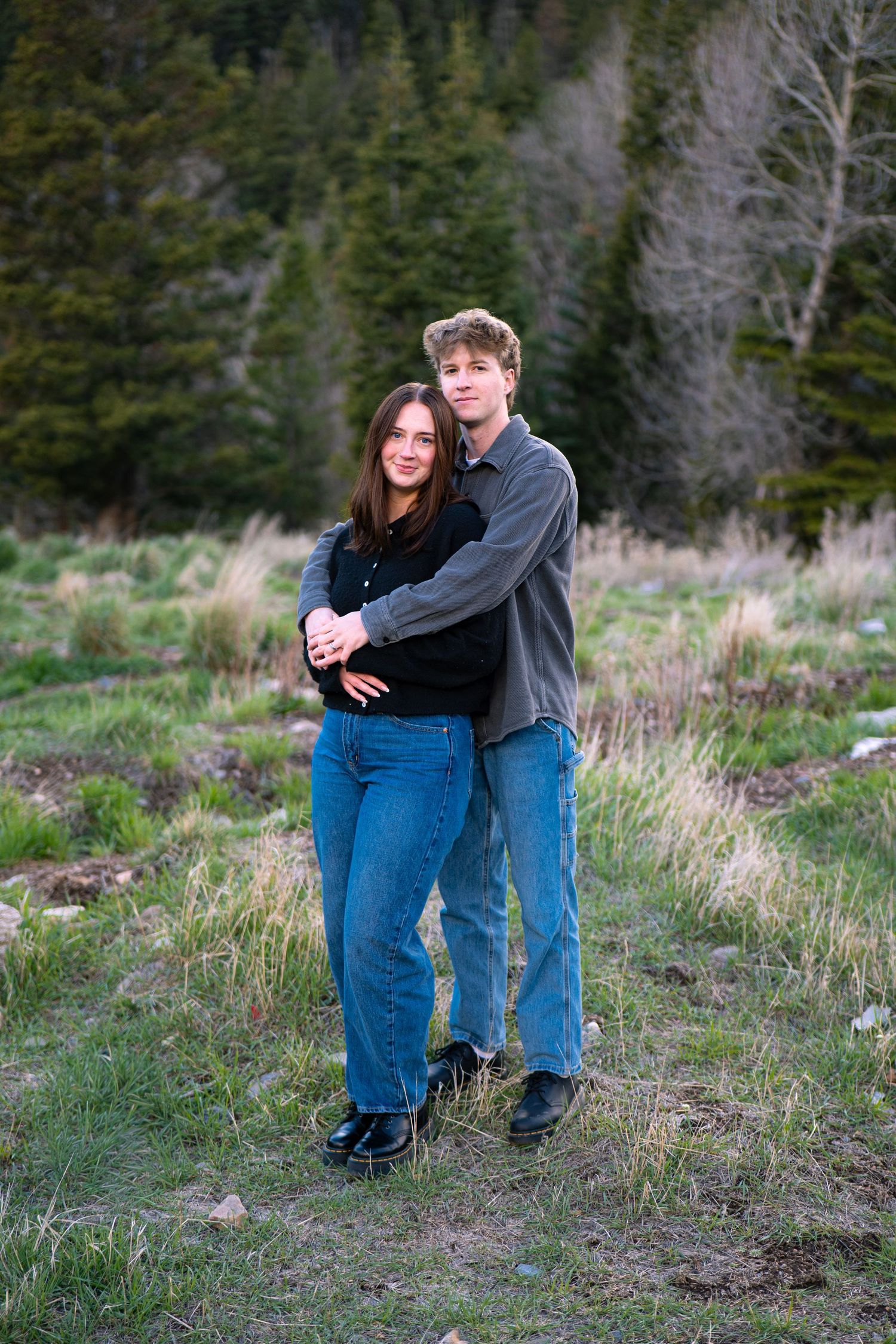 Young couple embracing outdoors in a green meadow with evergreen trees, wearing jeans and casual tops.