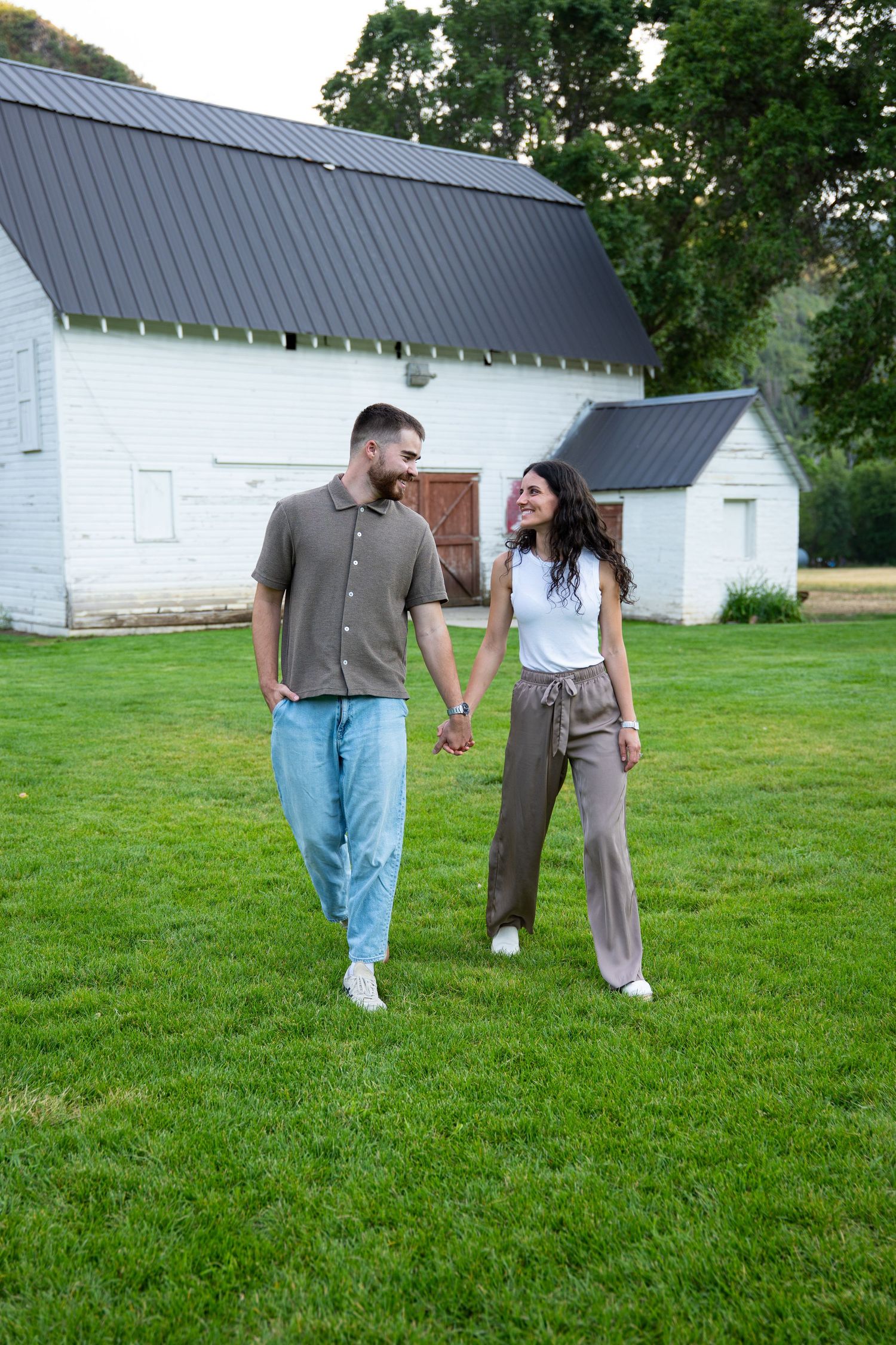 A couple holds hands and smiles while walking on green grass in front of a white barn.