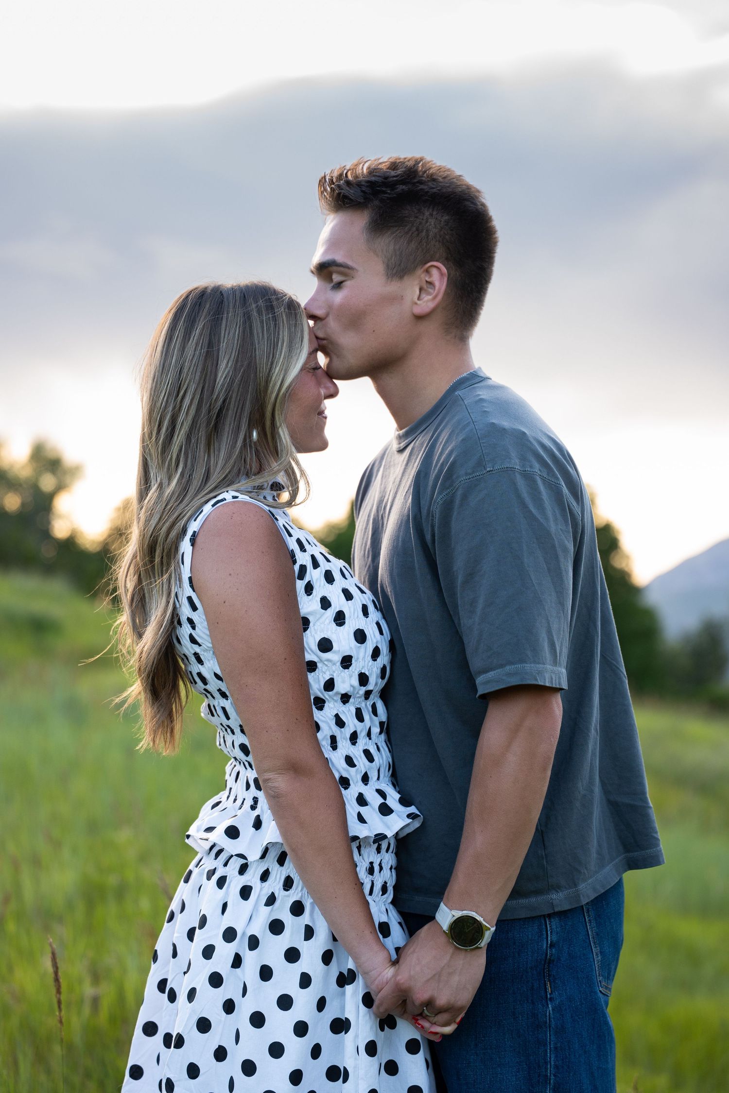 A young couple shares a tender kiss while holding hands in a green field at sunset.