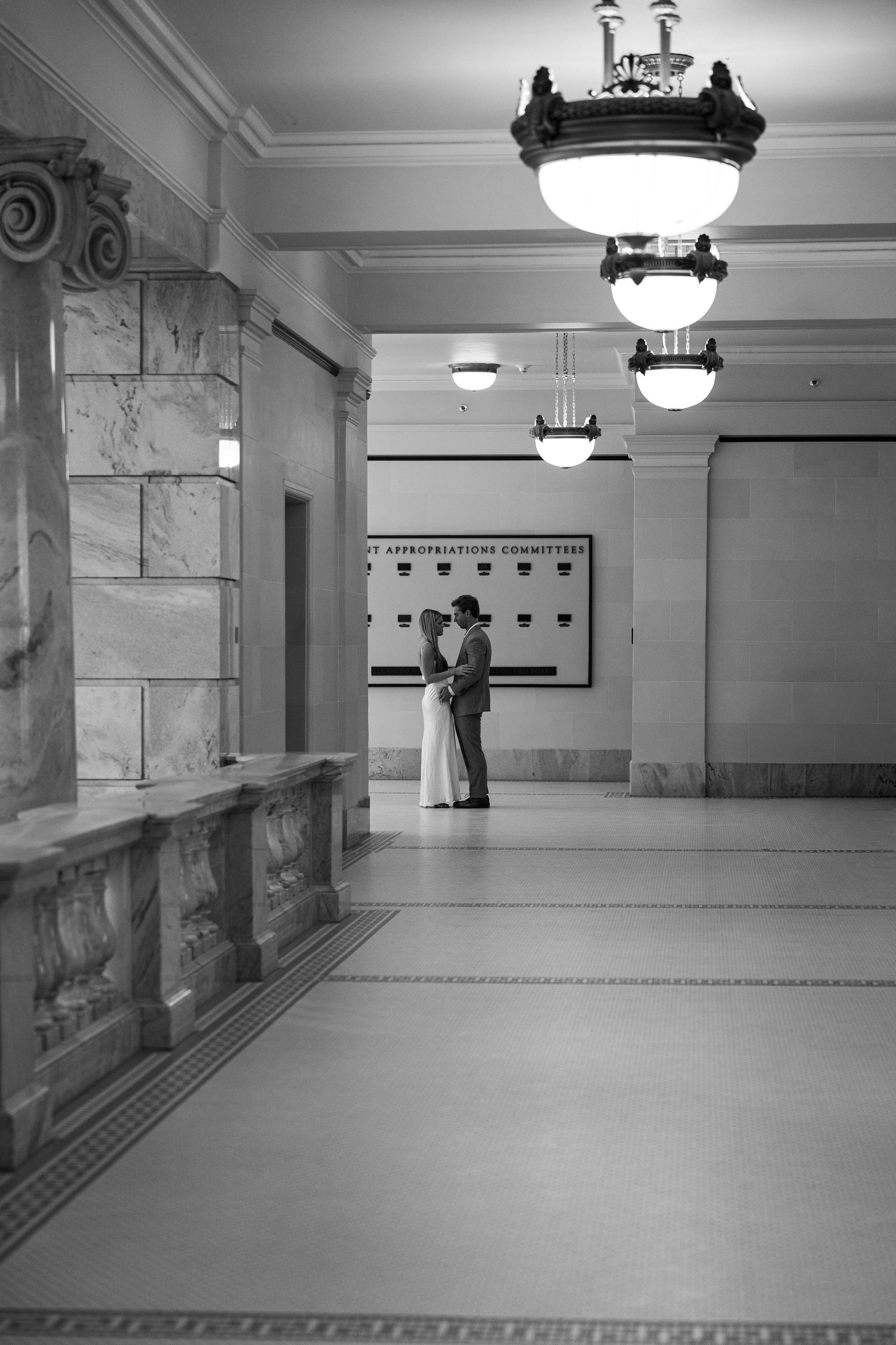 Couple sharing intimate moment in grand marble hallway with vintage chandeliers in black and white.