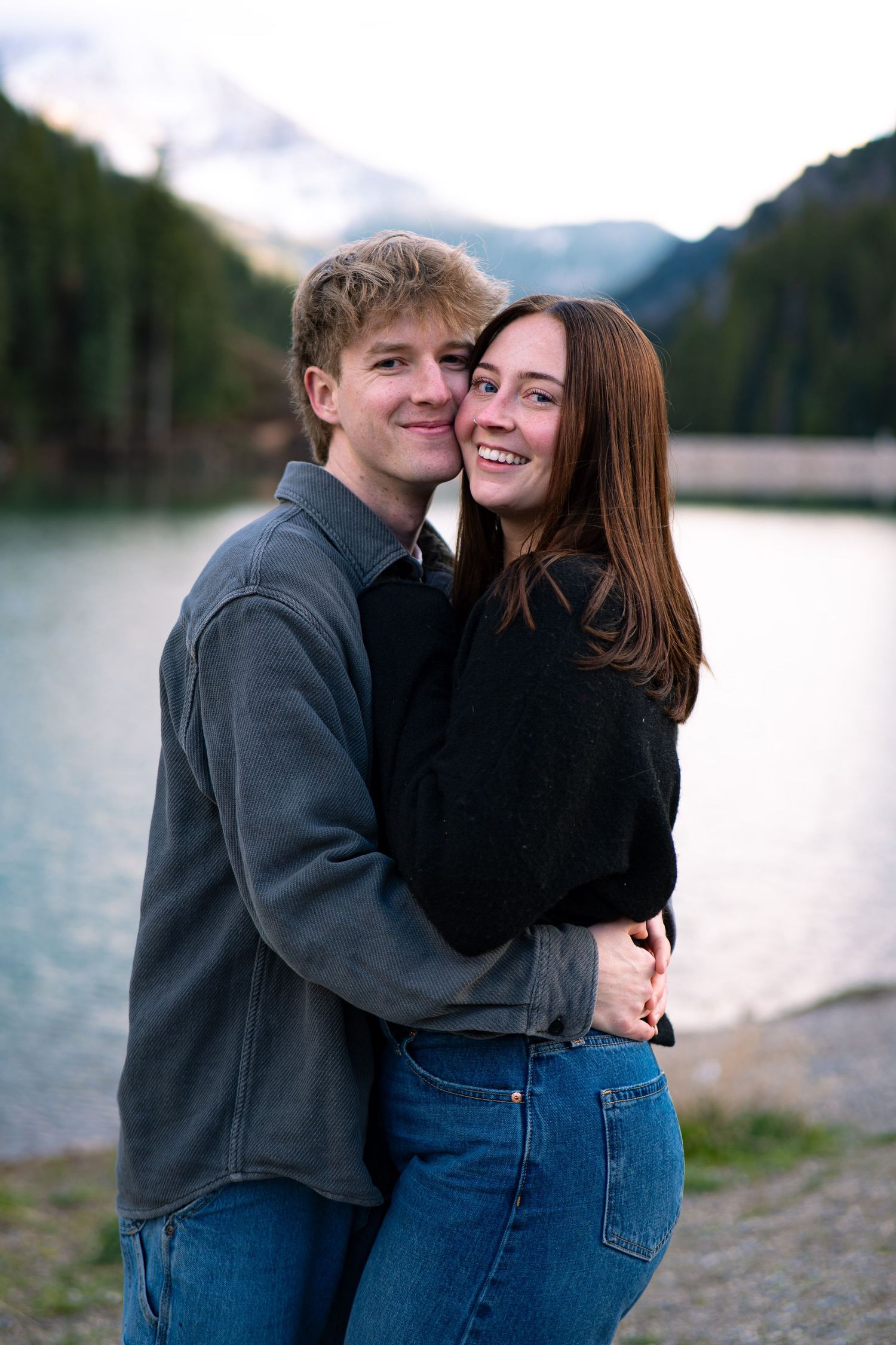 Happy young couple embracing and smiling by a scenic mountain lake with lush green trees in the background.