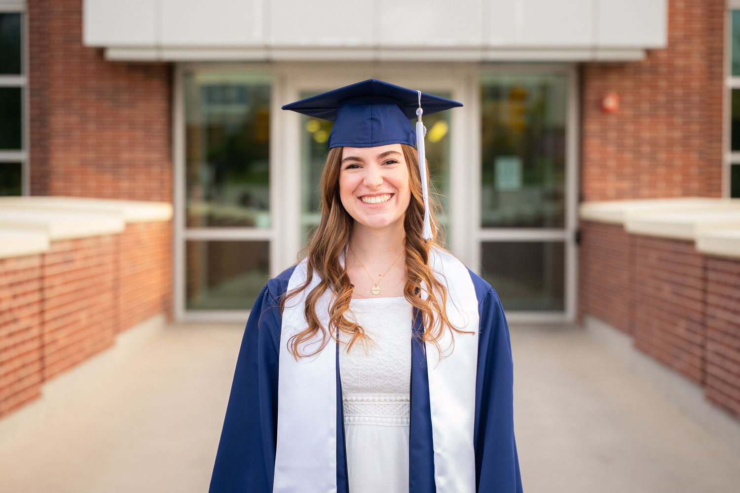 A smiling woman in a graduation gown and cap stands in front of a brick building.