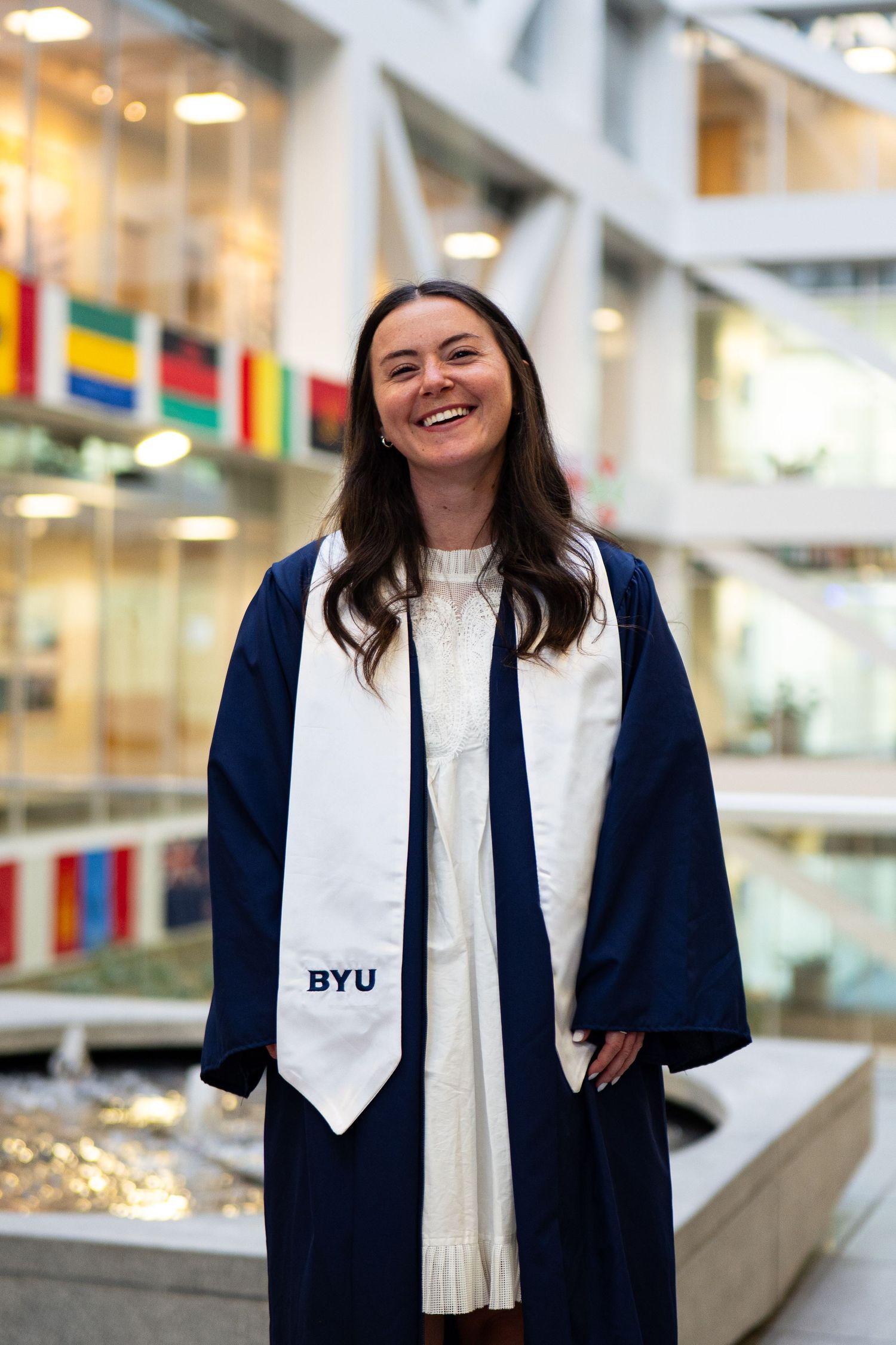 BYU graduate smiling in navy cap and gown with white BYU stole inside university building.