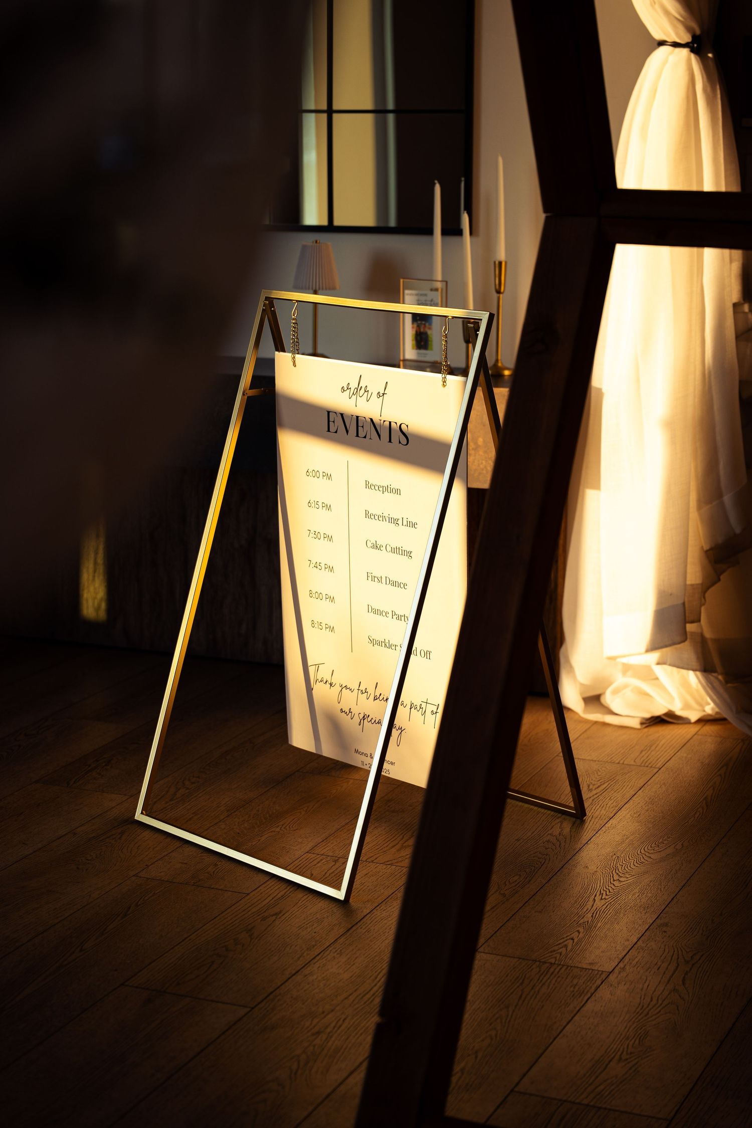 Elegant gold A-frame wedding event schedule sign displayed in a warmly lit venue with candles and draped curtains.