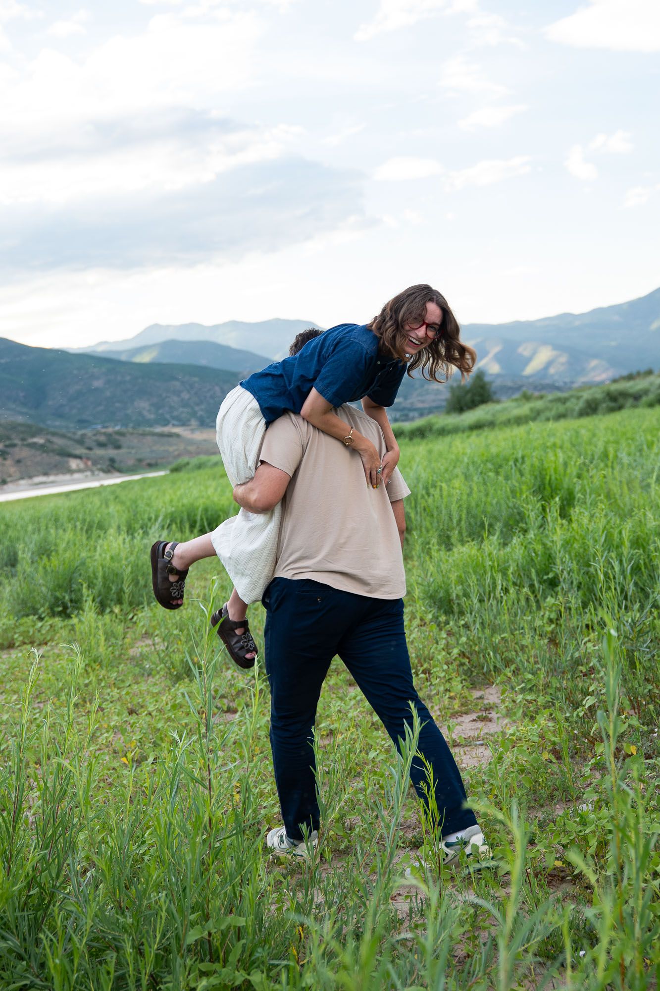 Couple playfully carries partner on back in lush green mountain meadow under cloudy sky.