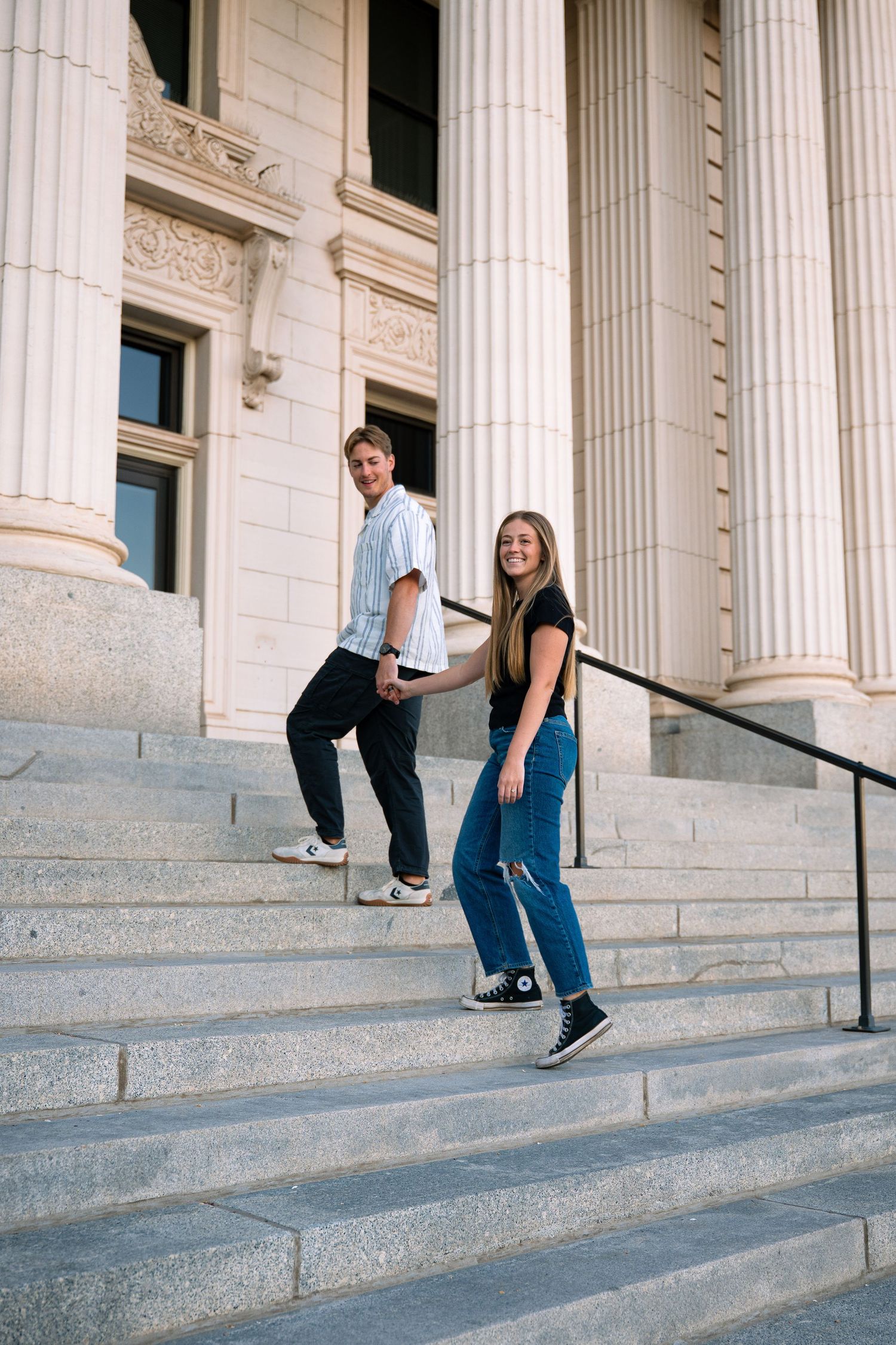 Young couple walking down stone steps of a neoclassical building, smiling, wearing casual clothes and sneakers.