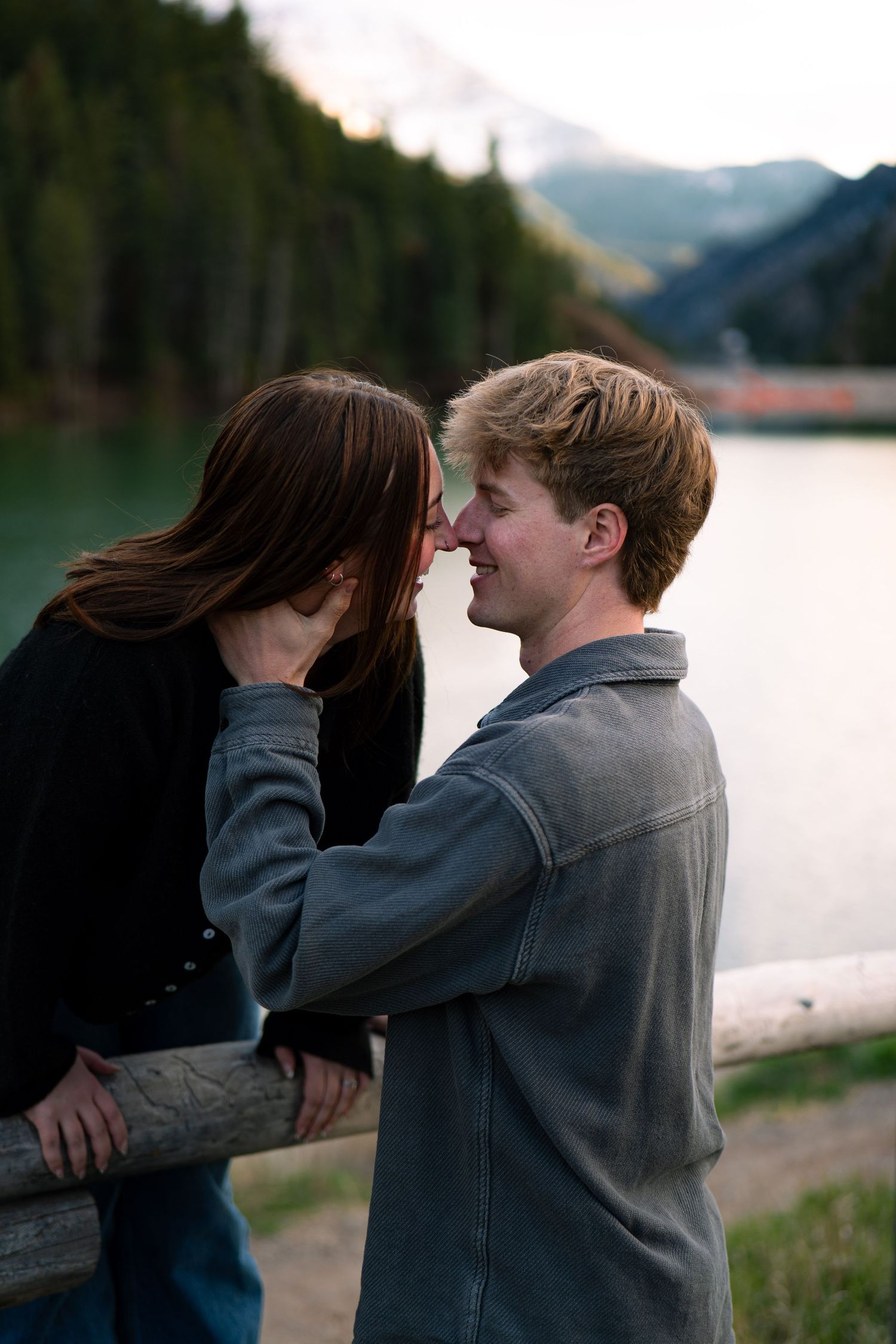 A young couple shares a romantic moment by a scenic mountain lake, nearly kissing while leaning on a wooden railing.