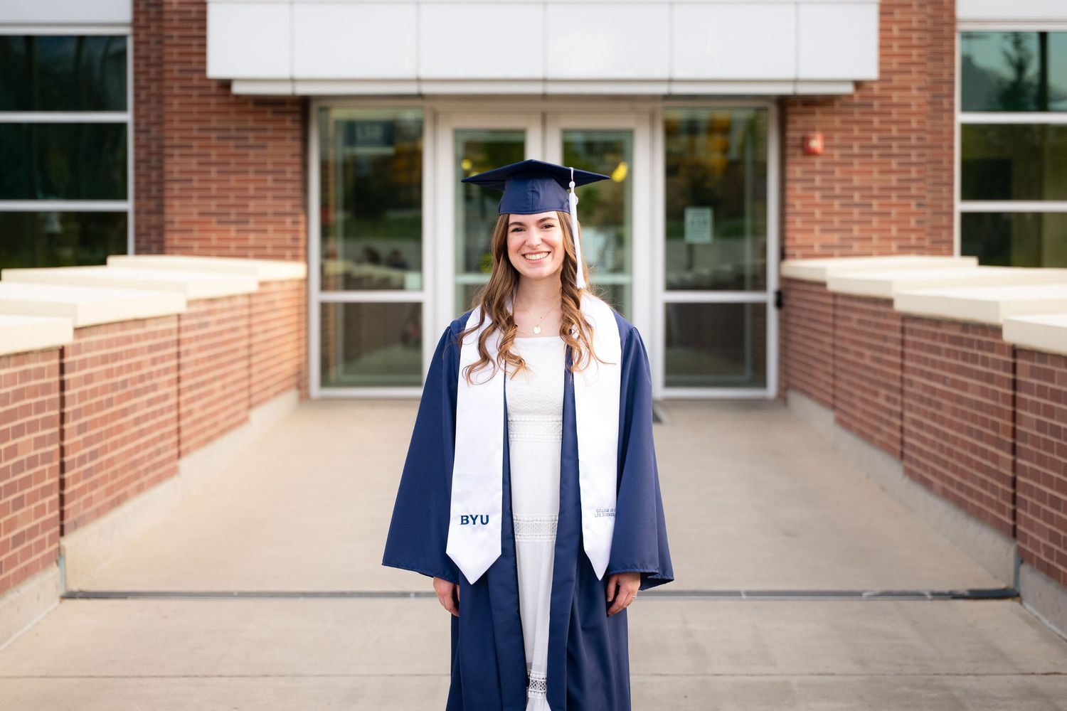 A smiling woman in a graduation gown and cap stands in front of a brick building.