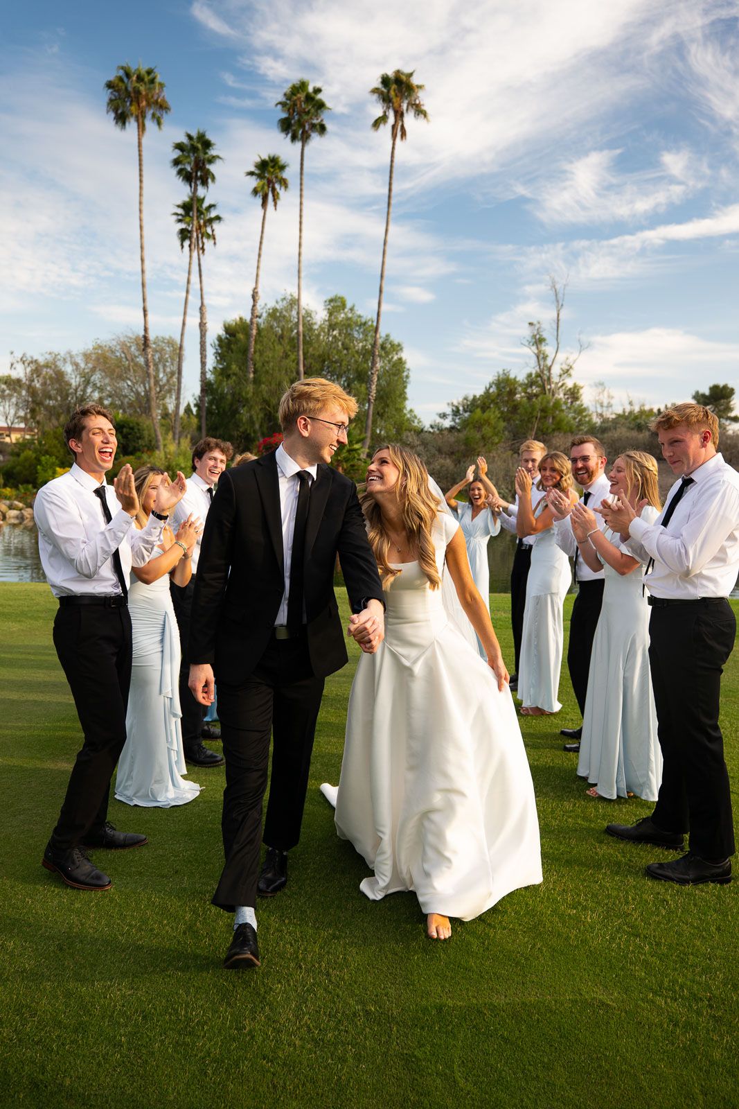 Bride and groom walk hand-in-hand through cheering wedding party on green lawn with palm trees.