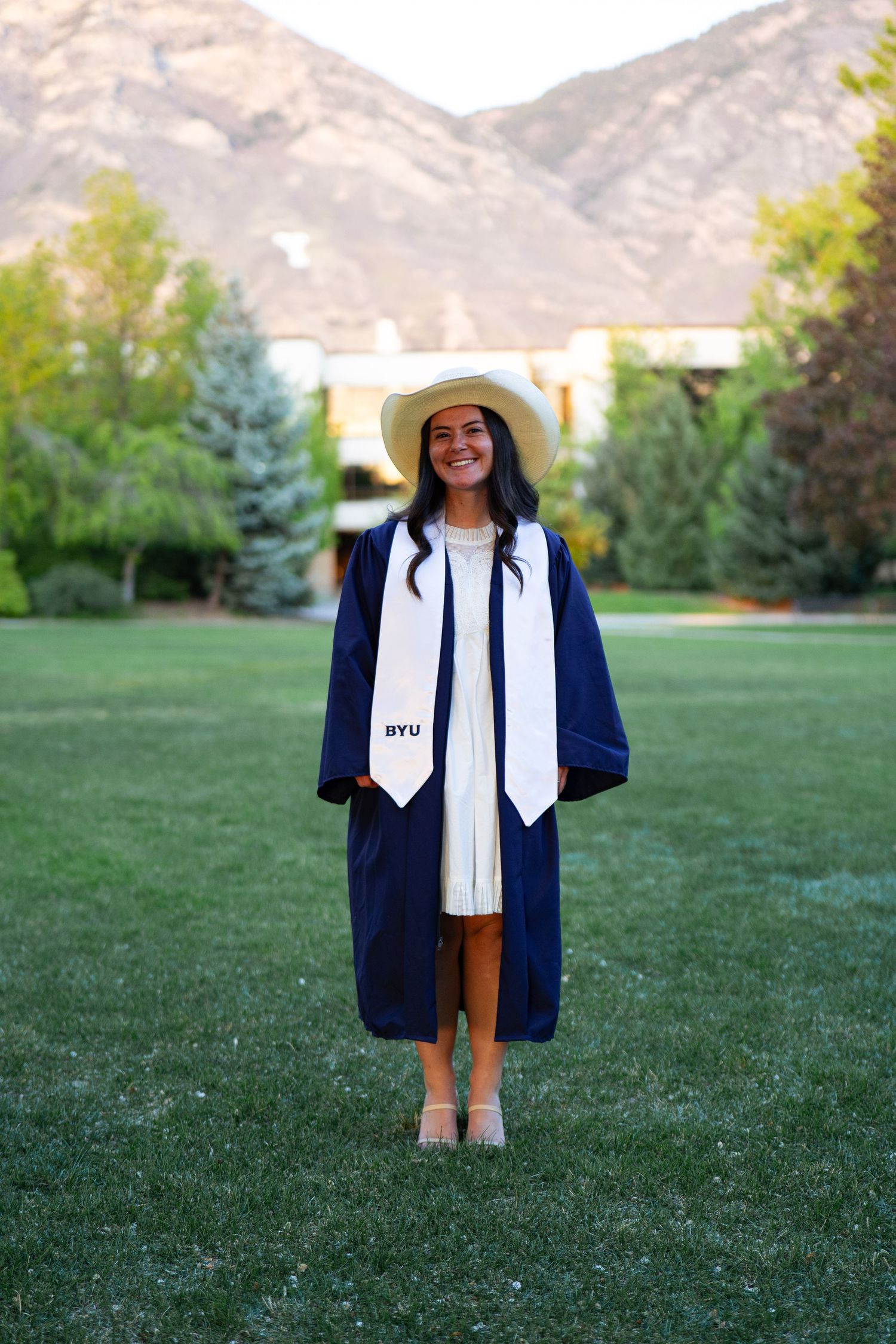 A smiling graduate in navy BYU cap and gown stands on green lawn with mountain backdrop.