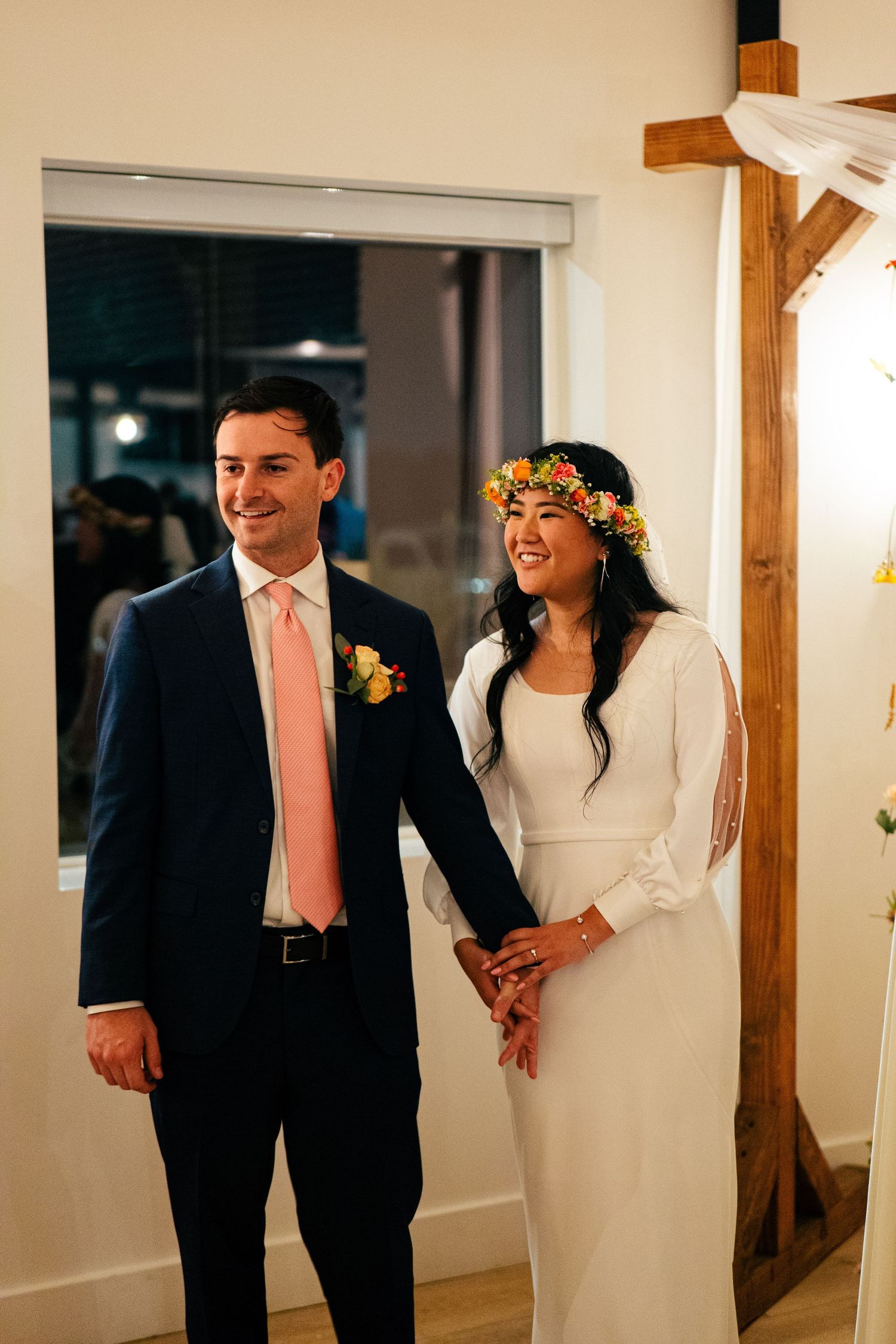 Happy newlyweds holding hands, bride wearing floral crown and white dress, groom in navy suit with pink tie.