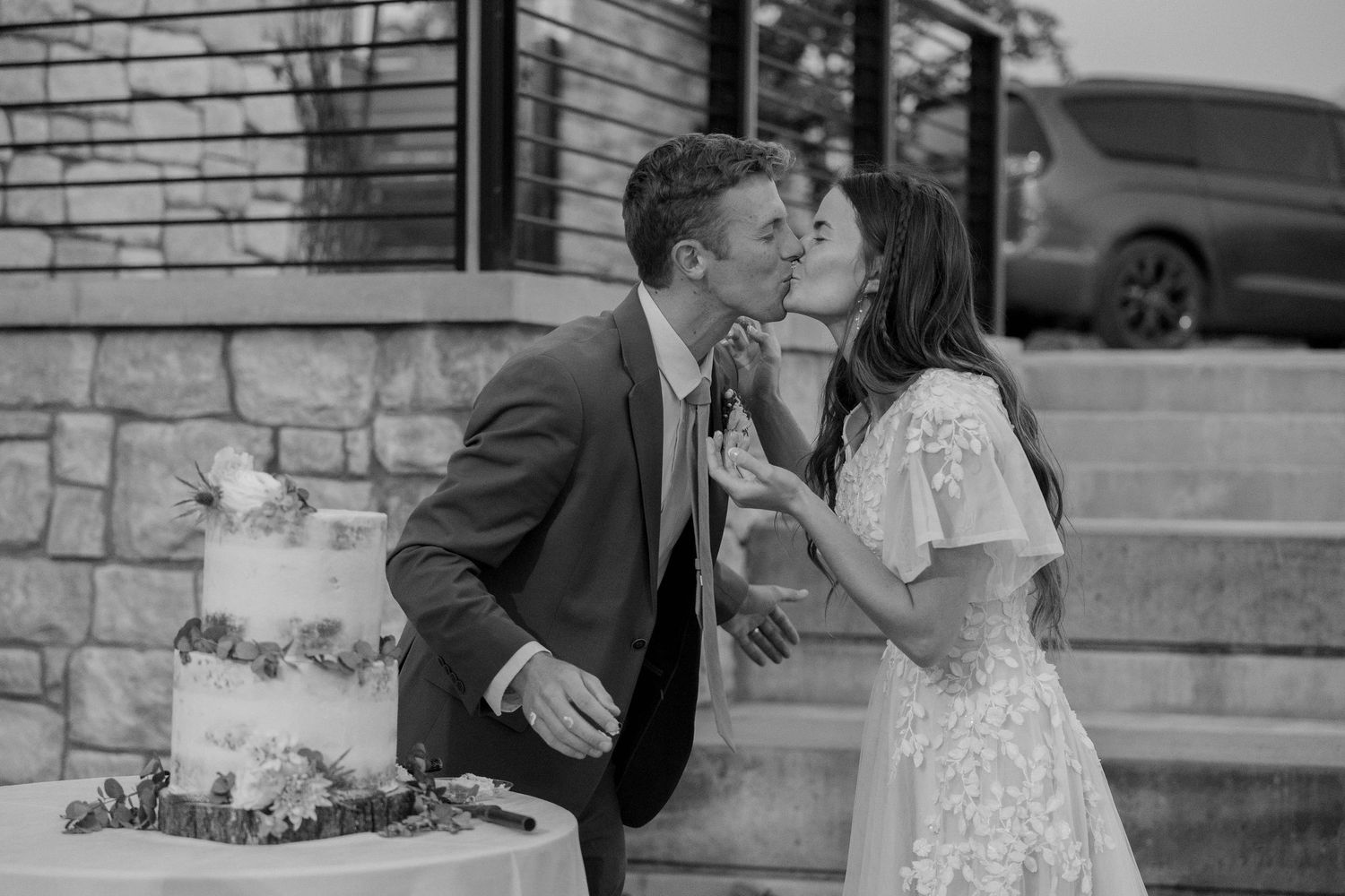 Newlyweds sharing a kiss while cutting their wedding cake in a black and white photo.