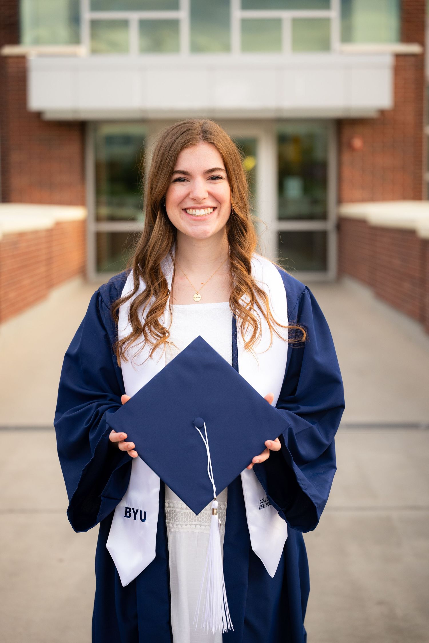 A smiling woman in a graduation gown and cap stands in front of a university building.