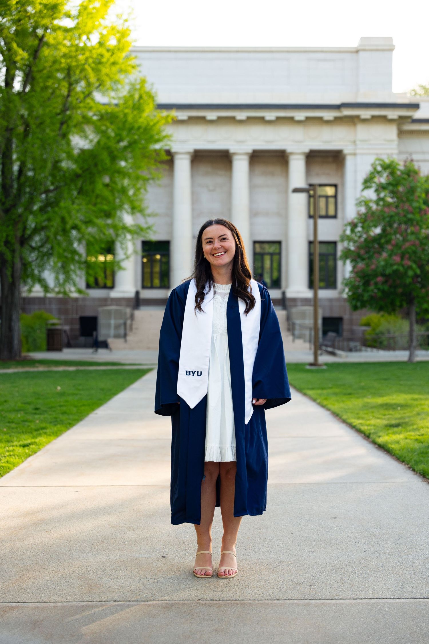 A smiling young woman in a navy BYU graduation gown and white stole stands on a campus walkway surrounded by green trees.