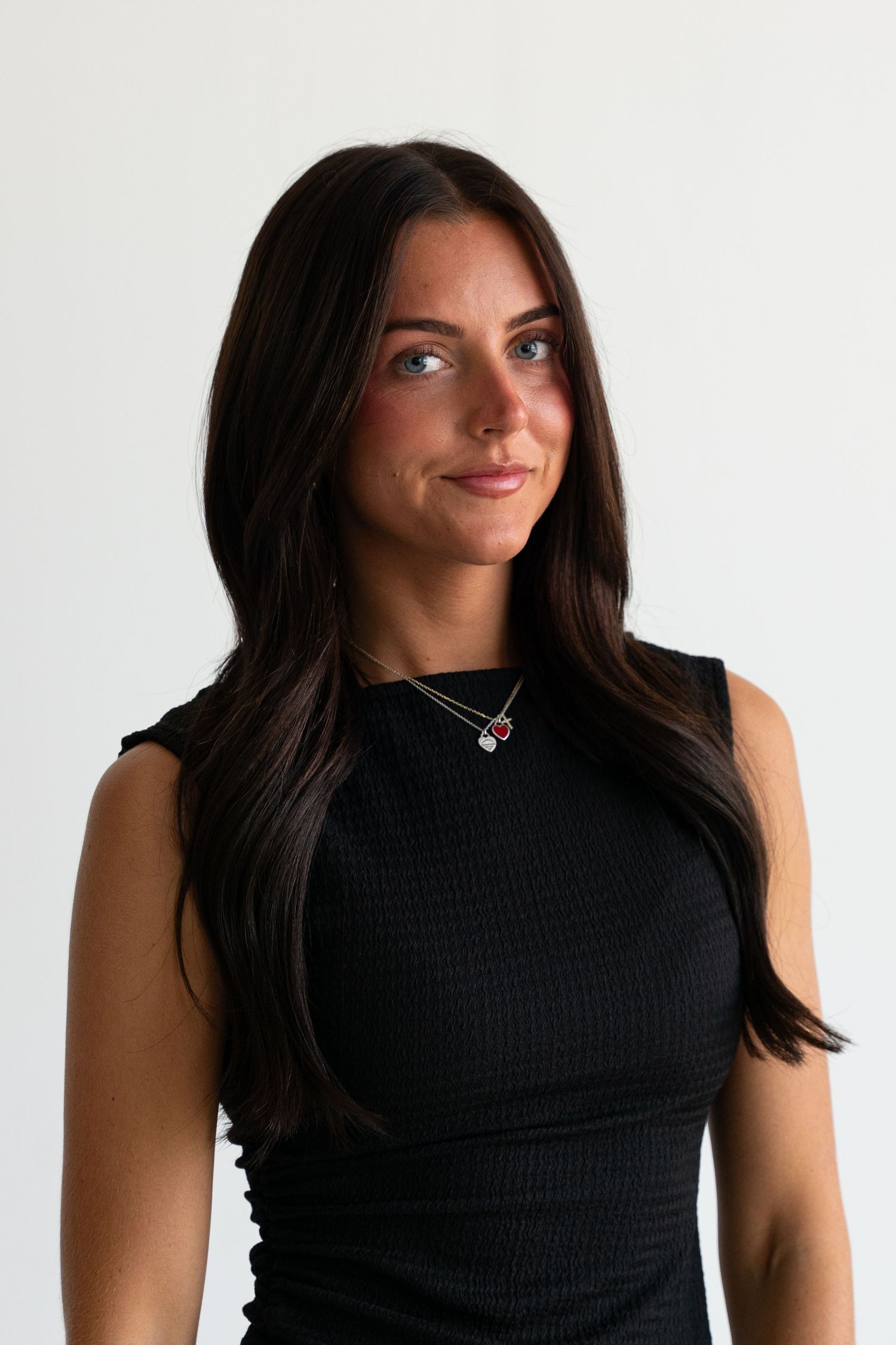 Young woman with long dark hair wearing a black sleeveless top and layered necklaces poses against white background.