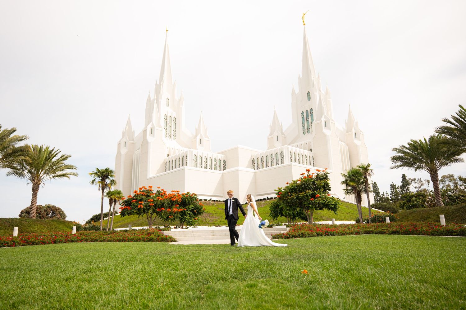 Bride and groom walking hand in hand on green lawn before a white LDS temple with tall spires.