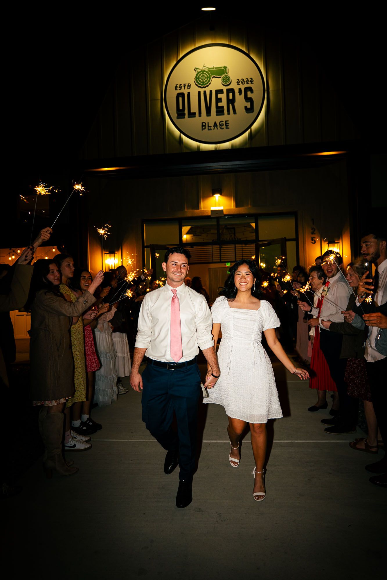 Couple exits wedding reception under sparklers at Oliver's Place venue at night.