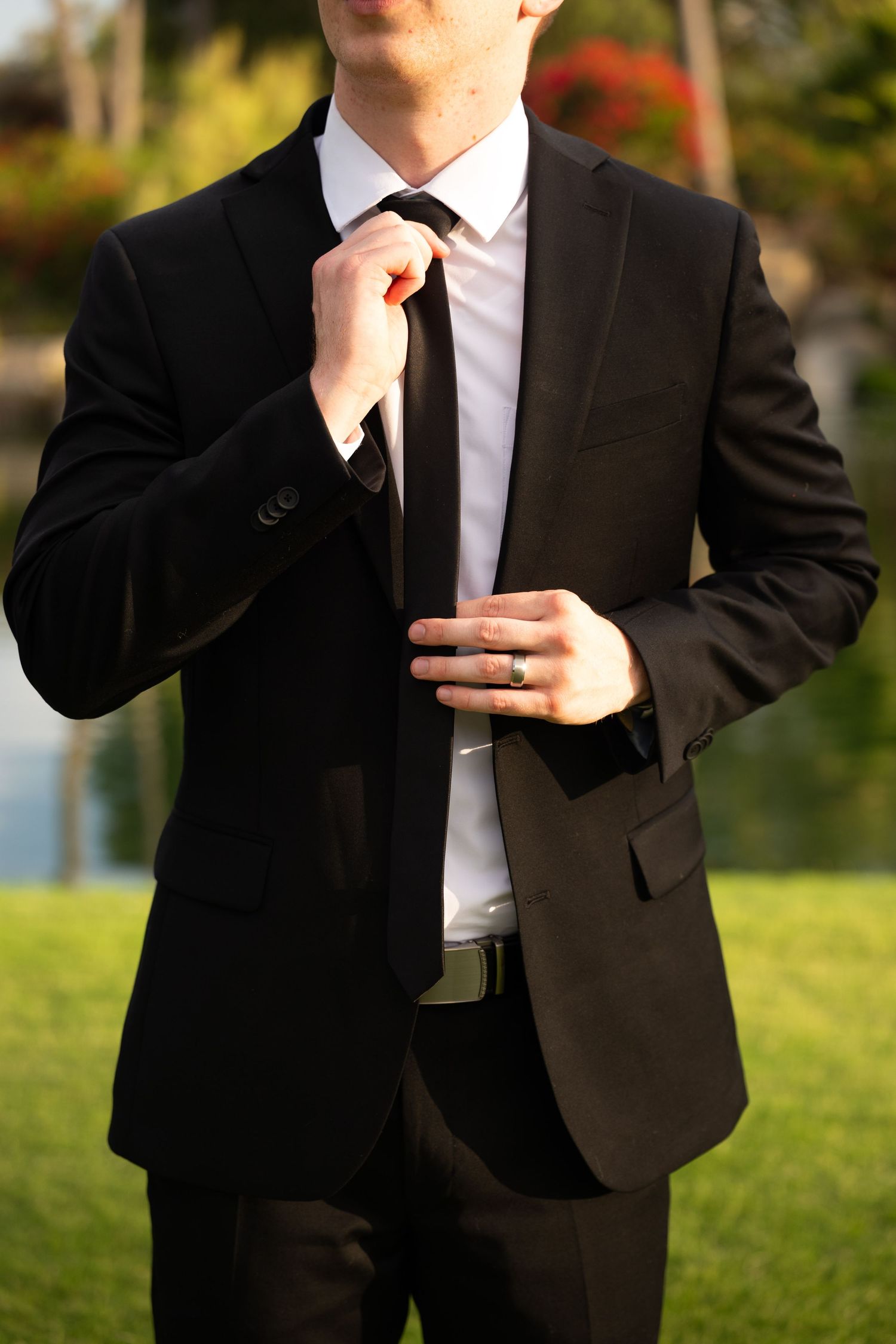 A groom in a black suit and white shirt adjusts his black tie outdoors, wearing a wedding ring.