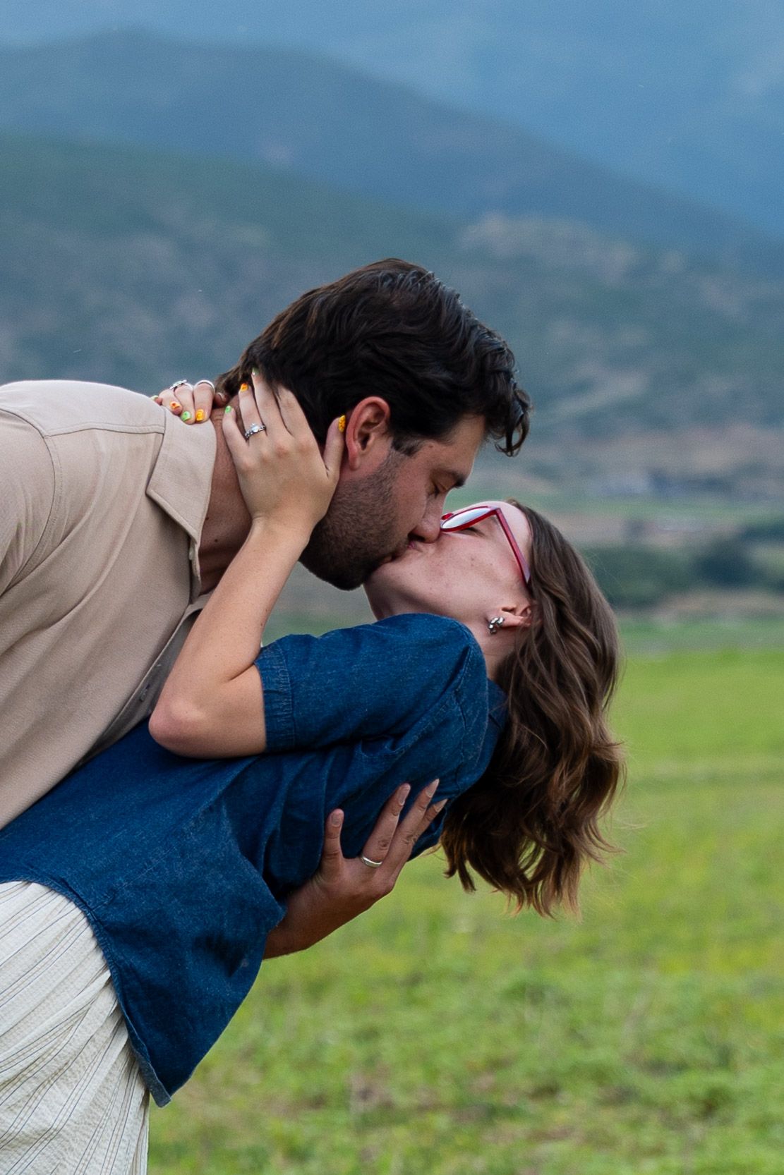 Couple sharing a romantic dip kiss outdoors with scenic green mountains in the background.
