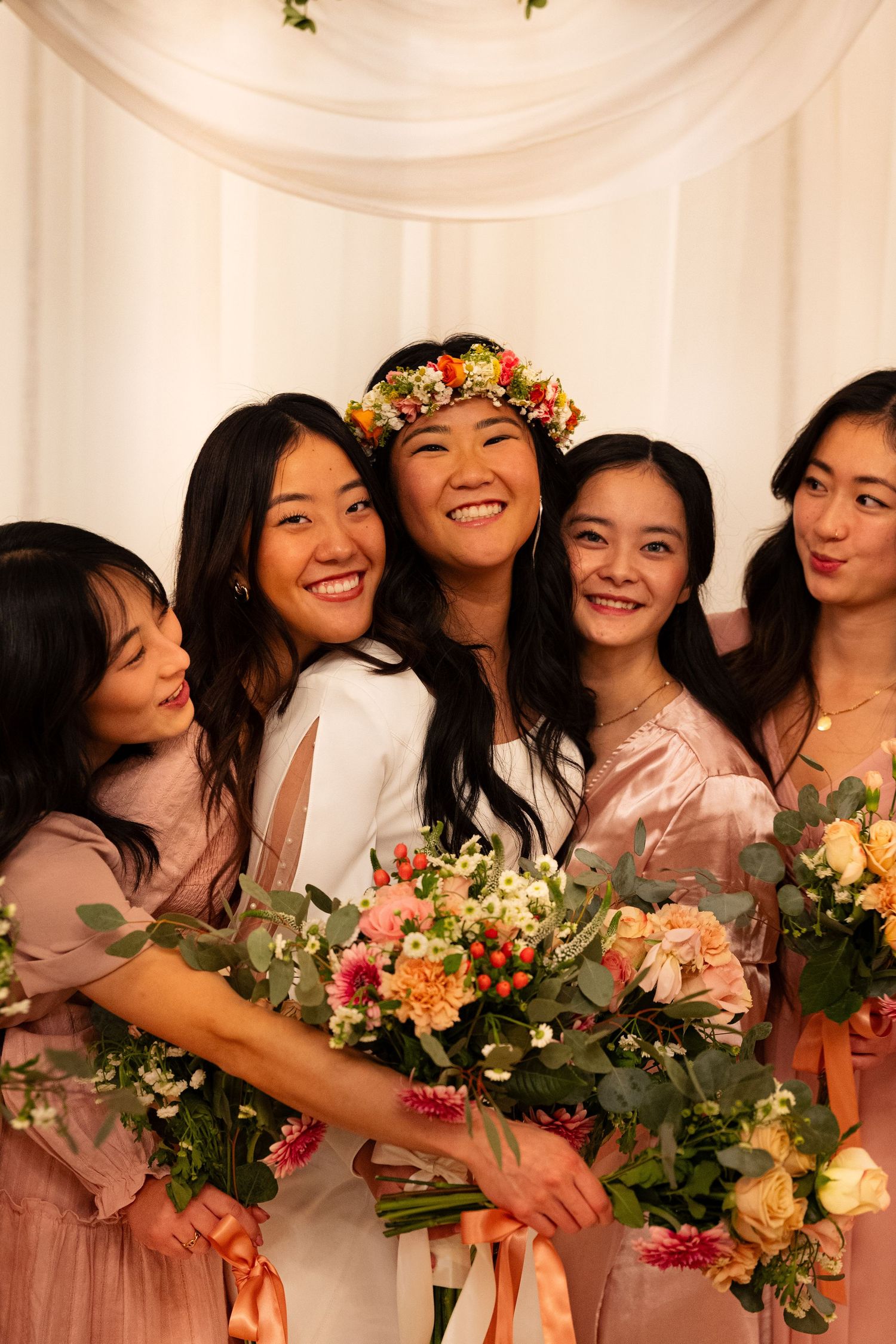 Smiling bride wearing floral crown poses with bridesmaids in pink dresses holding colorful bouquets at wedding ceremony.