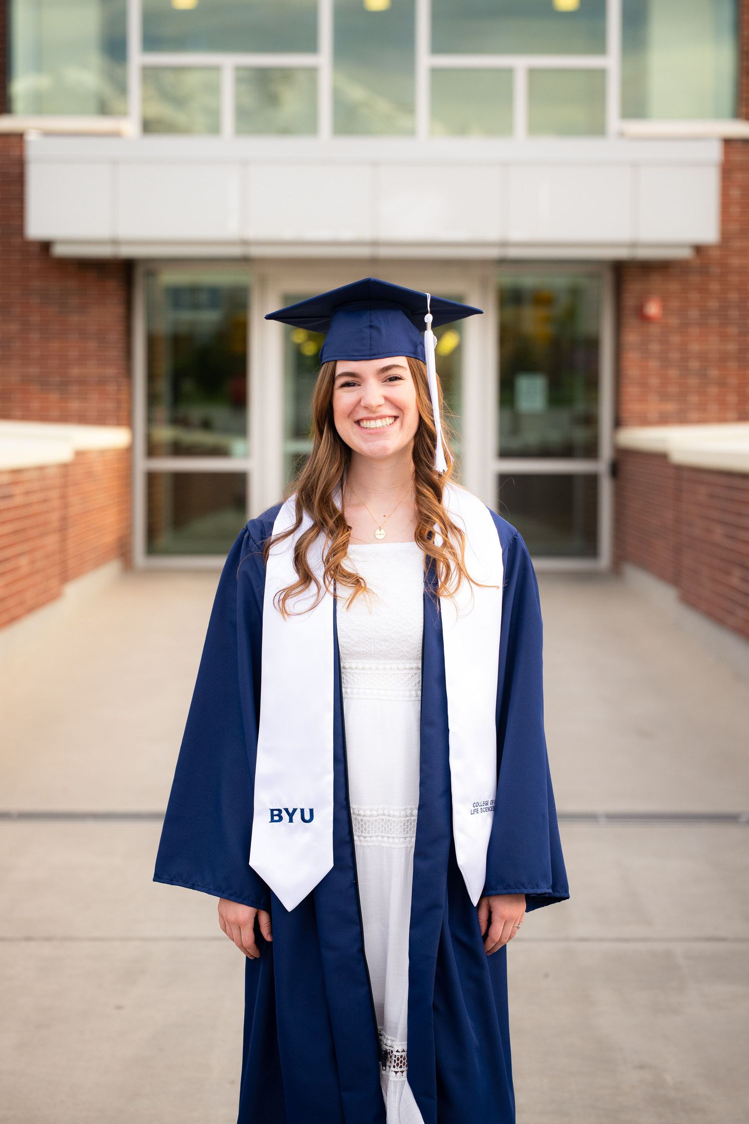 A young woman with long curly brown hair smiles while wearing a white dress and blue graduation gown in front of a building.