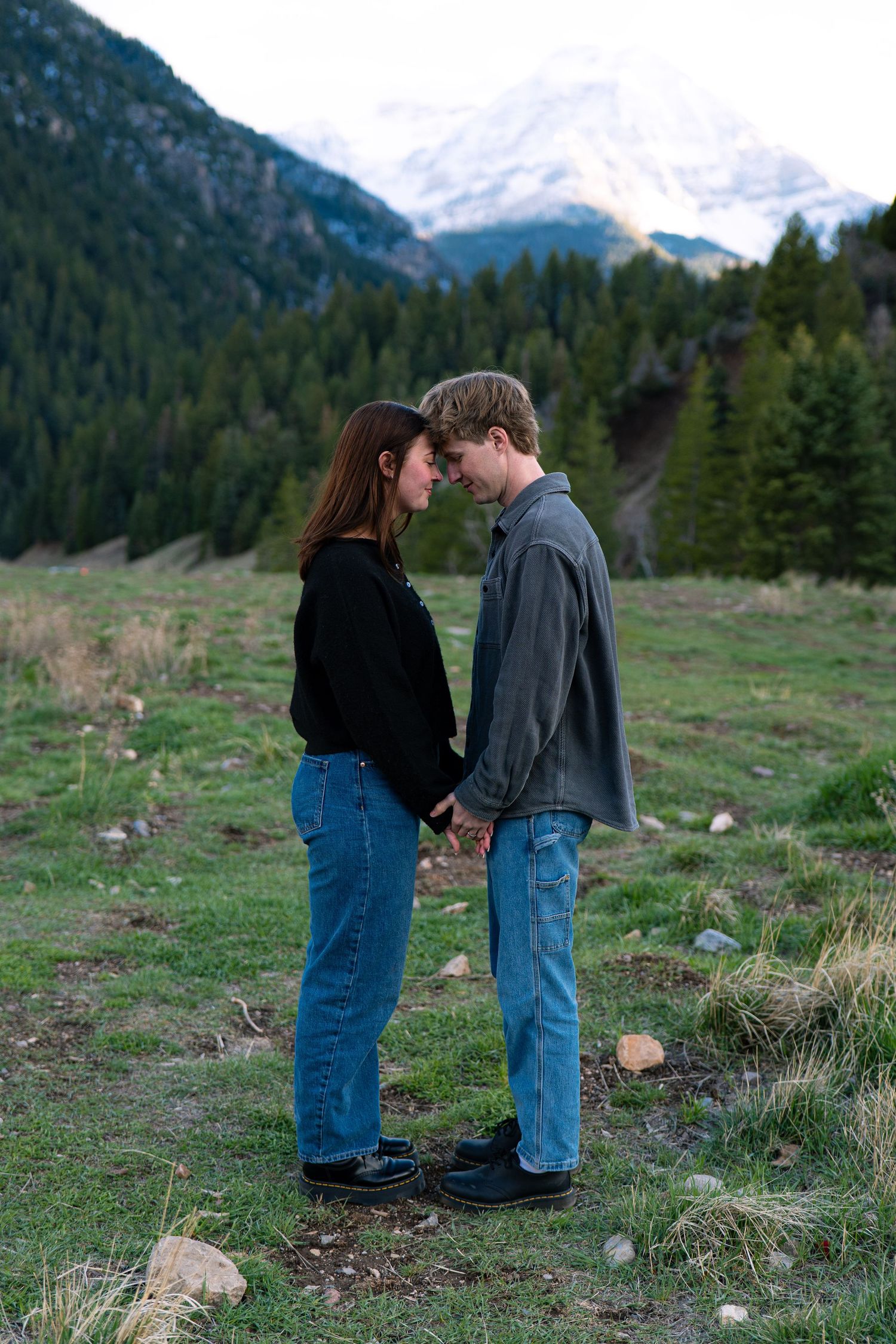 Couple holding hands facing each other in a mountain meadow with snow-capped peaks and pine trees in background.