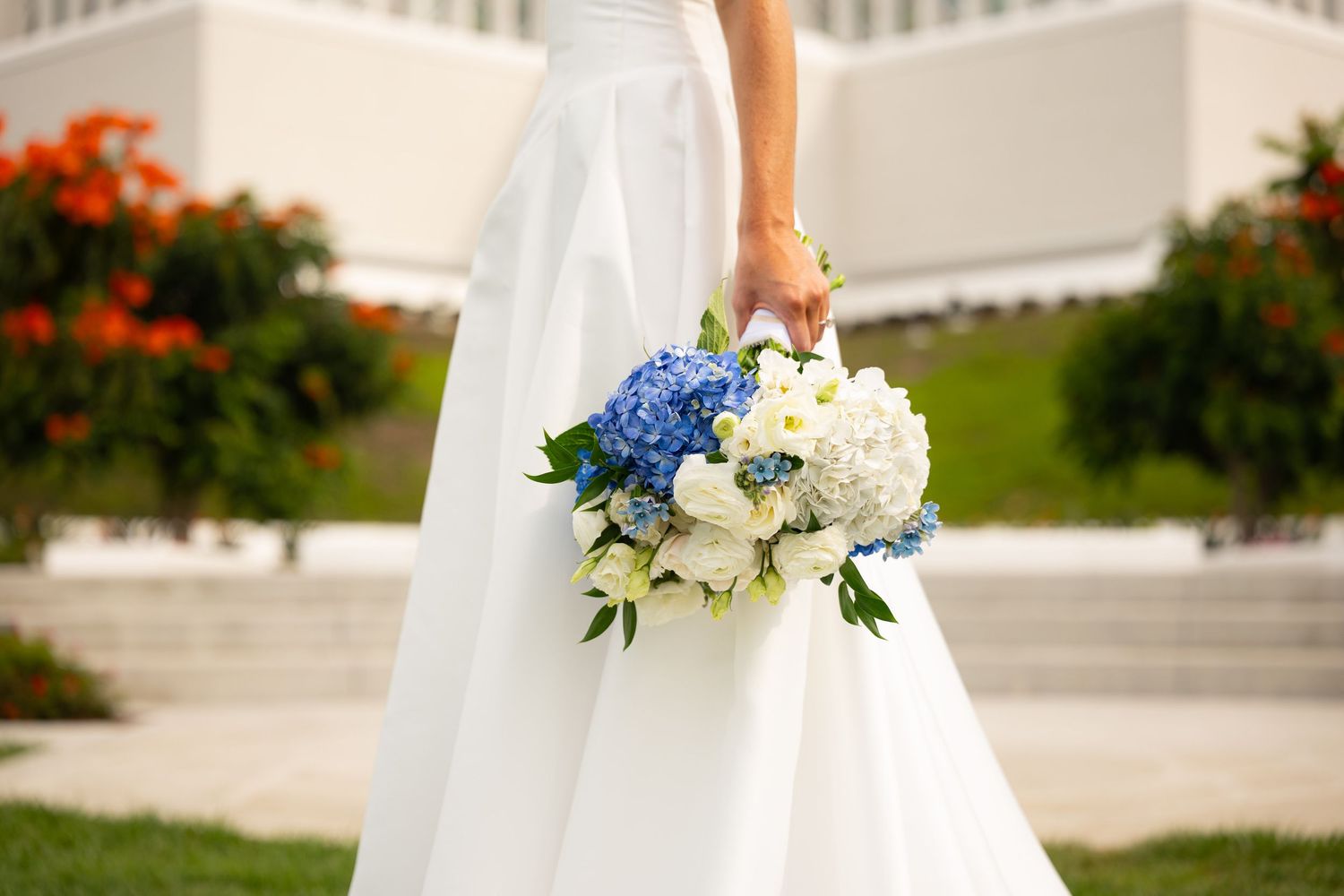 Bride in white gown holding blue hydrangea and white peony wedding bouquet outdoors near blooming red flowers.