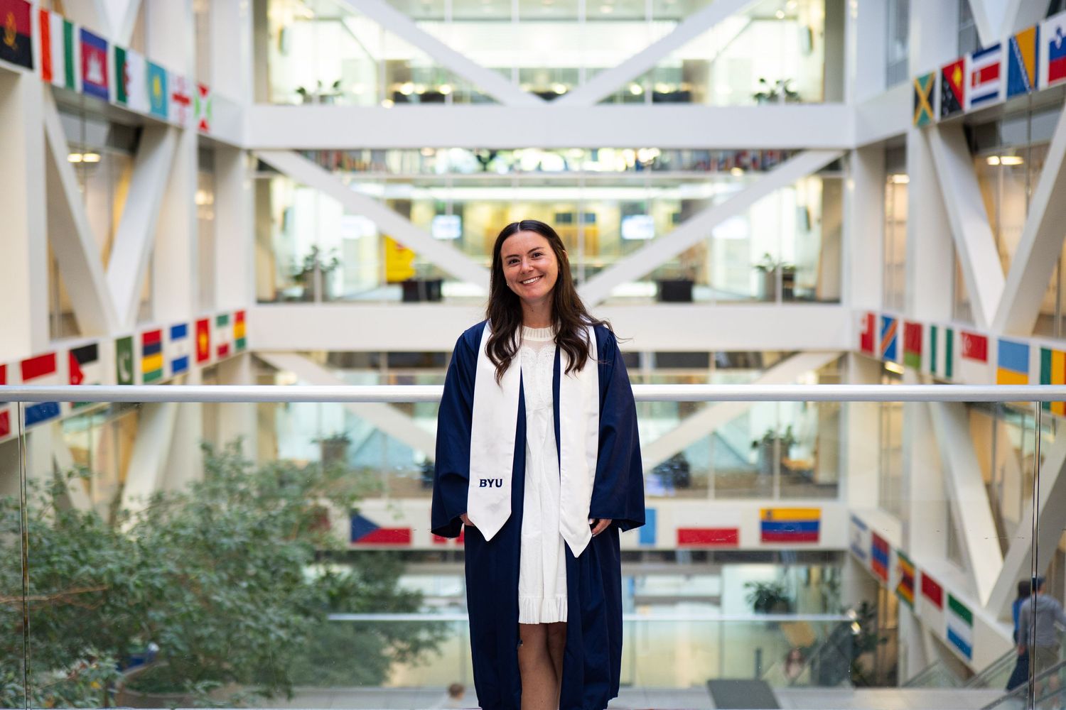 A smiling graduate in navy cap and gown stands in a bright atrium decorated with international flags.